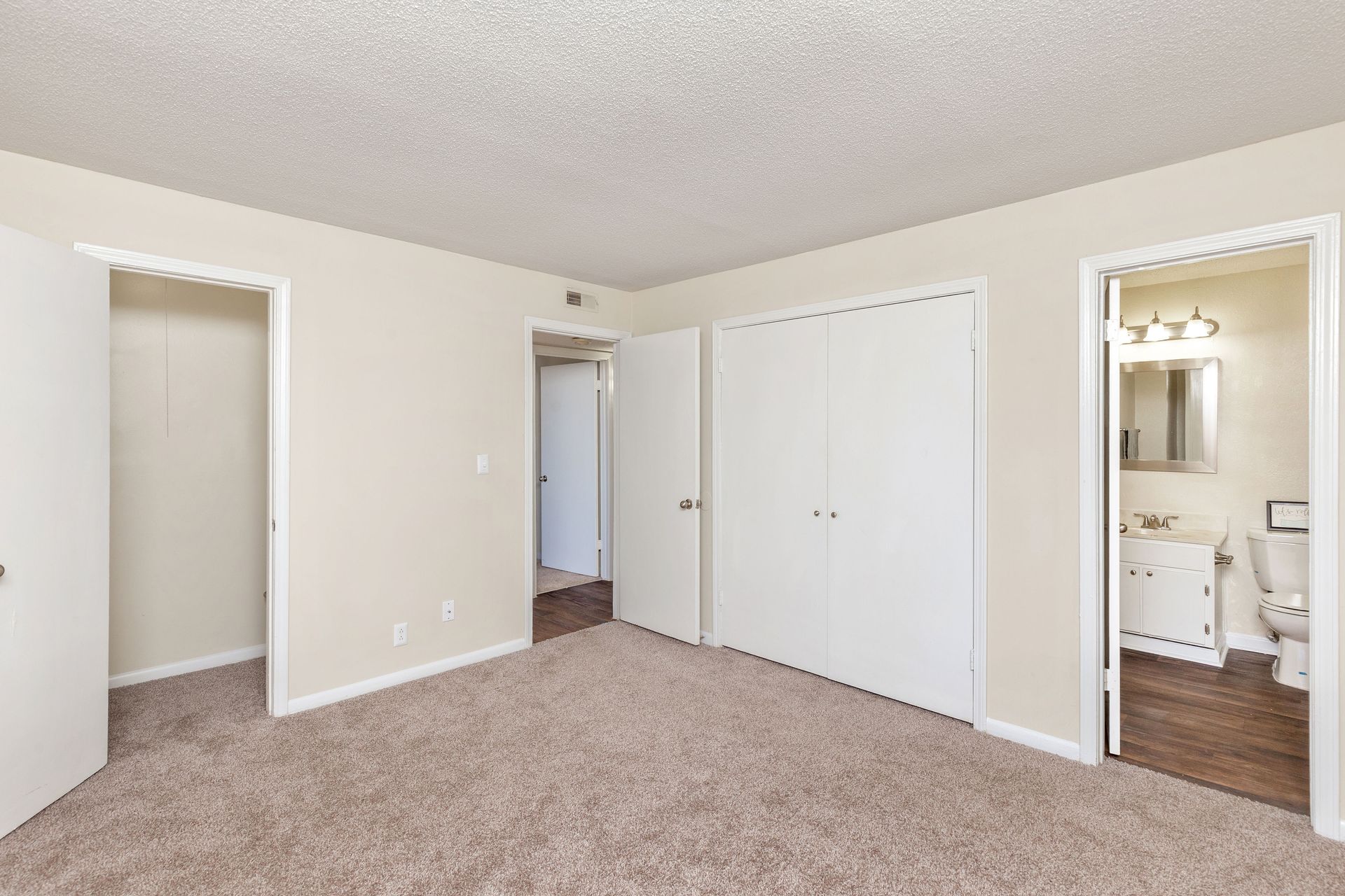 An empty bedroom with beige carpet, light walls, a closet, a hallway entrance, and an attached bathroom.