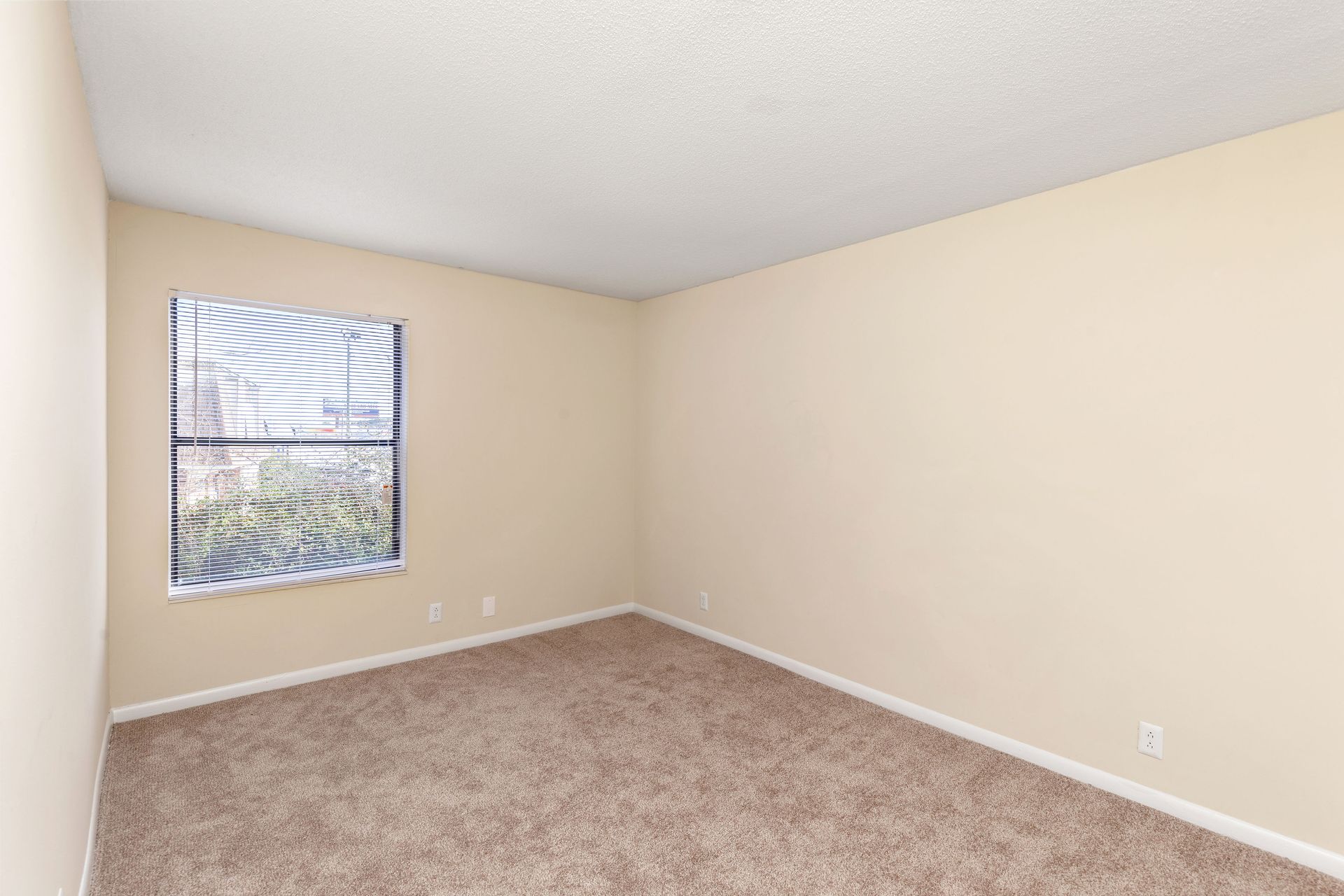 An empty bedroom with beige walls, beige carpet, a white ceiling, and a single window with closed horizontal blinds.