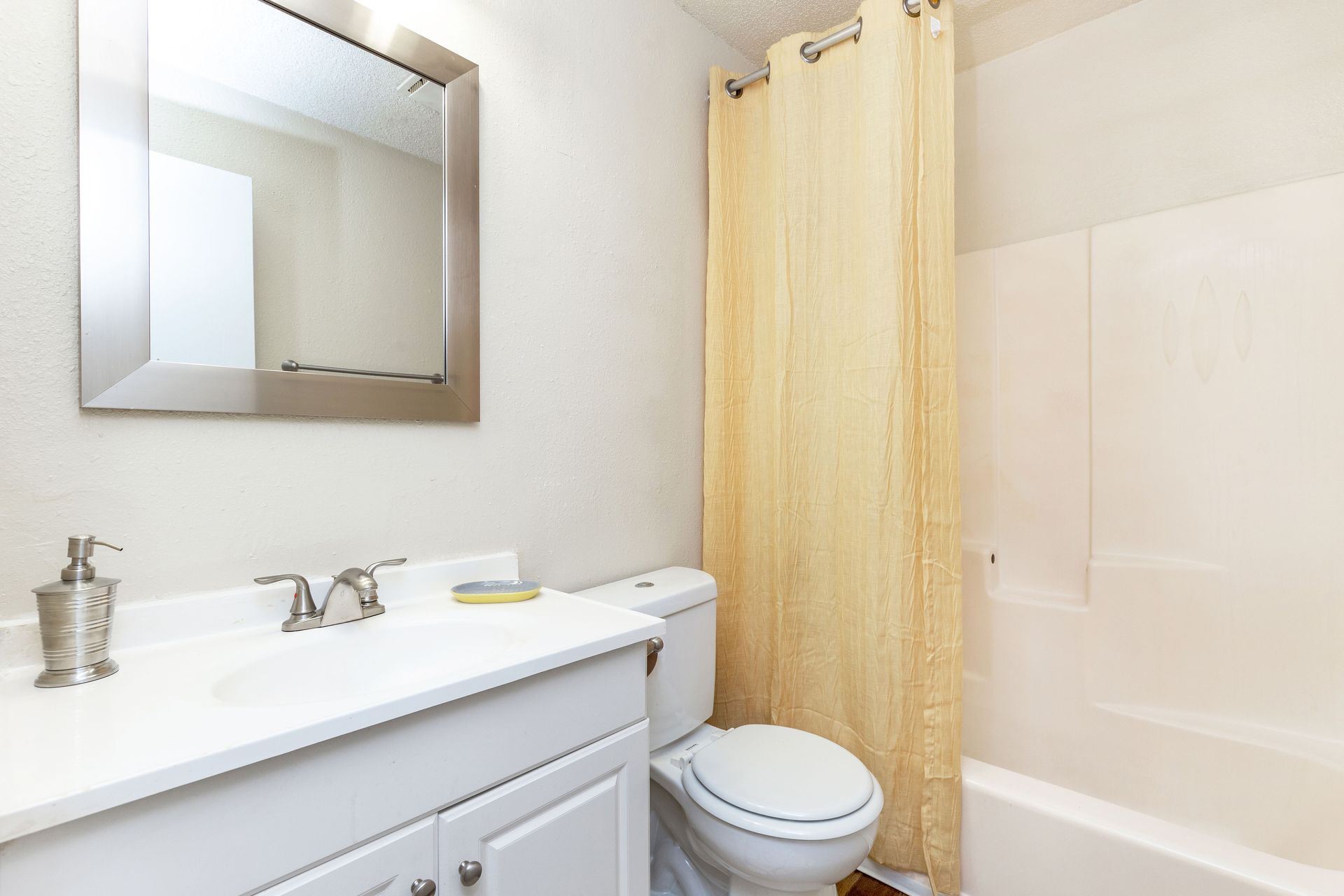 A modern bathroom featuring a white vanity with a silver faucet, a framed mirror, a toilet, and a bathtub with a tan curtain.