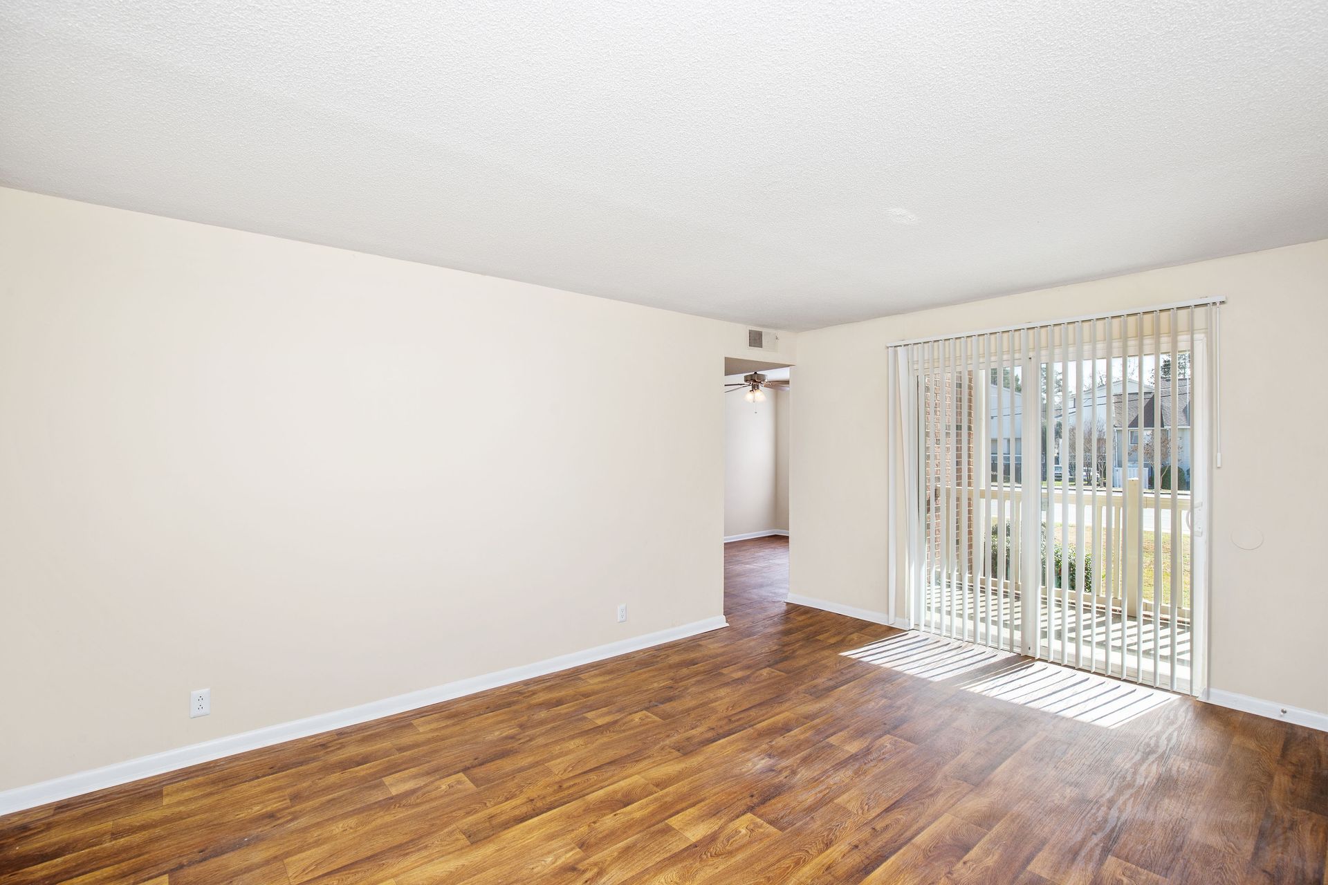 An empty room with light beige walls, wood-look flooring, a popcorn ceiling, and a sliding glass door with vertical blinds.