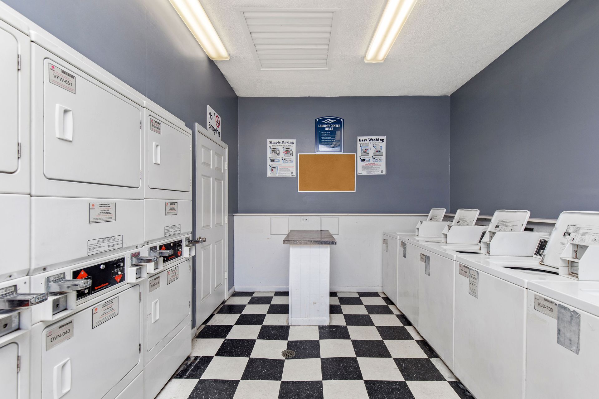 A brightly lit laundry room with a checkerboard floor, white washers and dryers along side walls, and a center table.