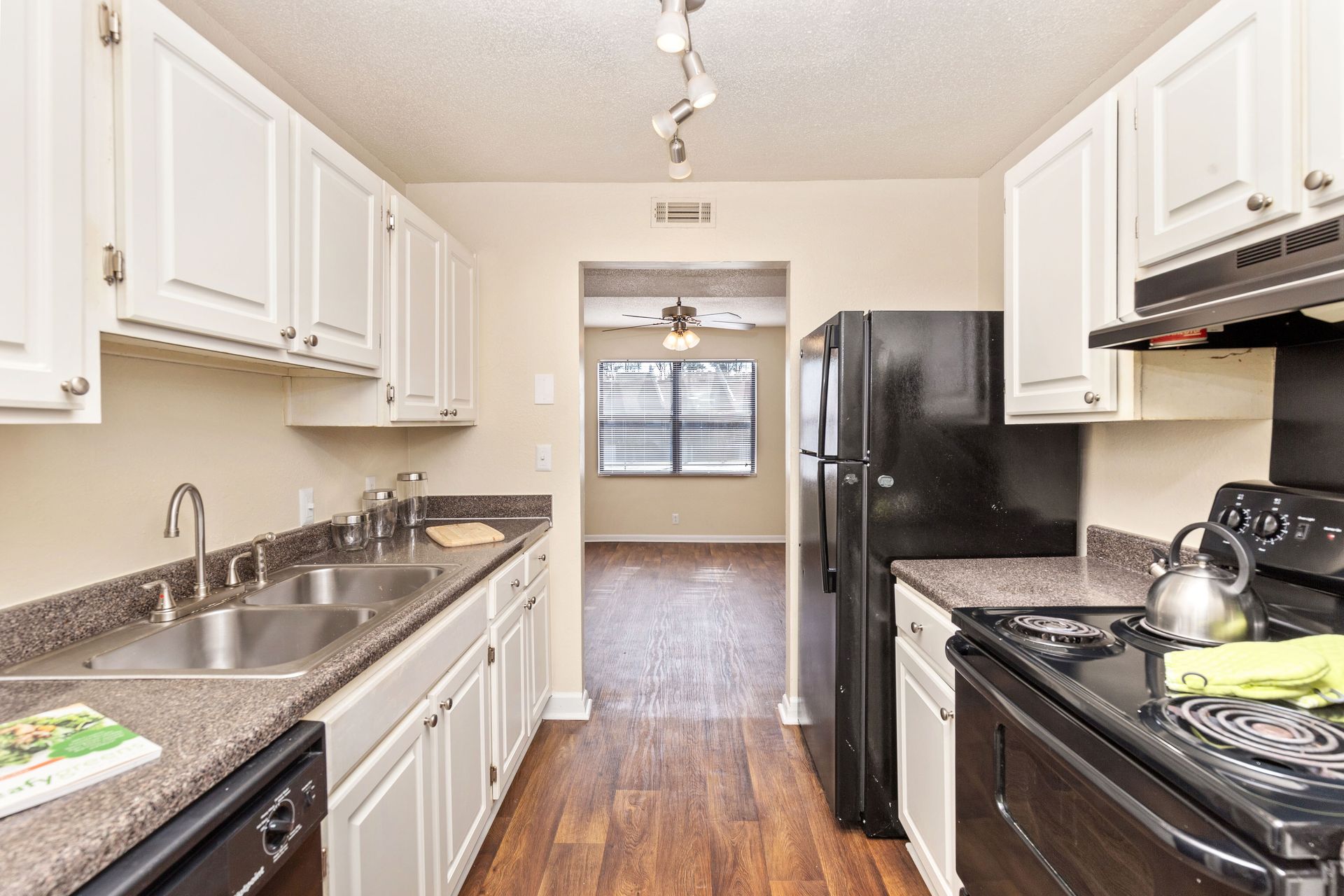 A galley kitchen with white cabinets, dark granite-look countertops, black appliances, and wood-look flooring.