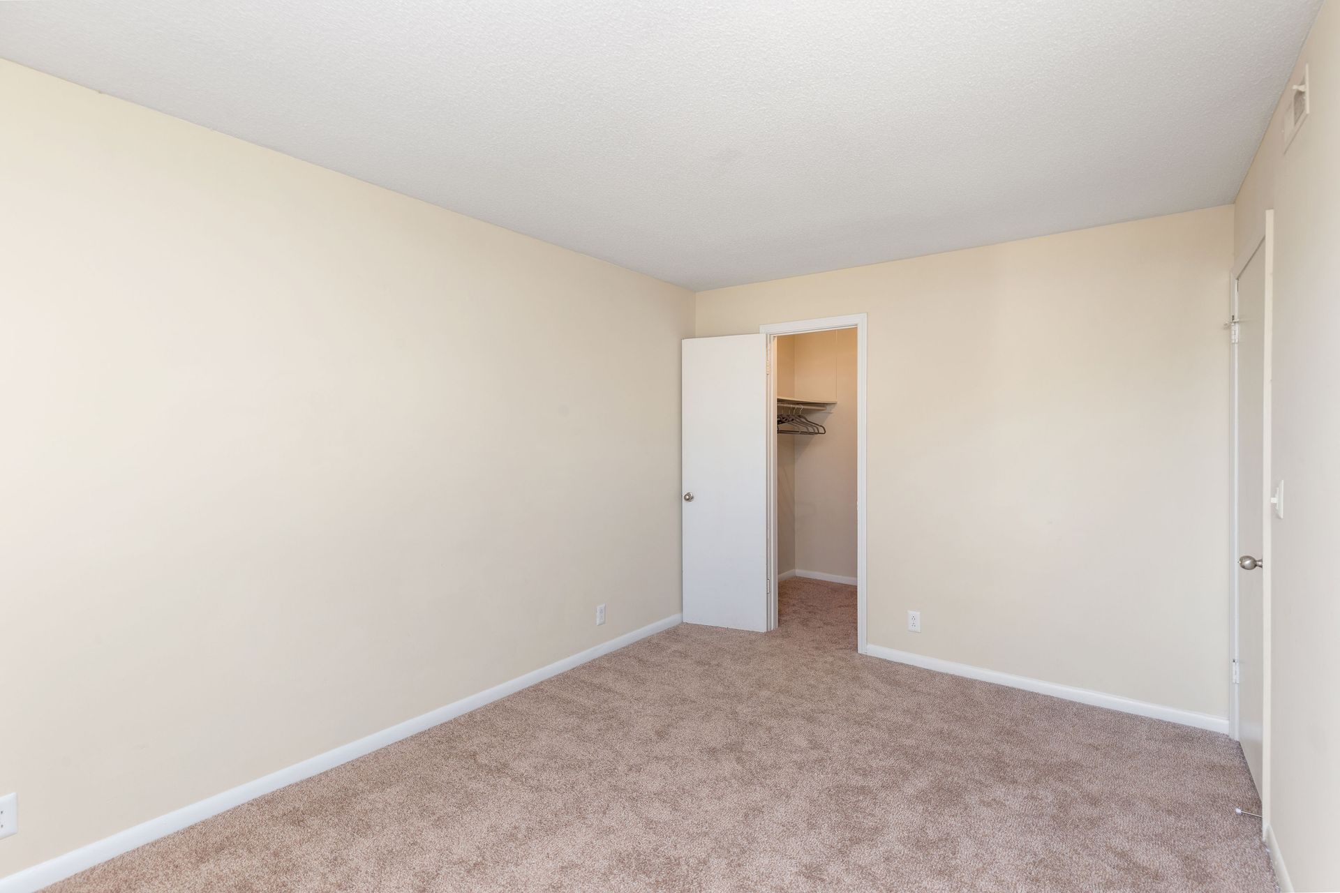 Empty bedroom with beige walls, brown carpet, an open closet door, and a closed door on the right wall.