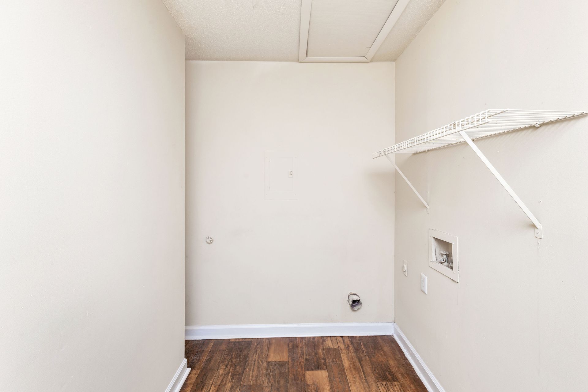 A laundry closet with beige walls, a wire shelf, and hookups for a washer and dryer over a hardwood floor.