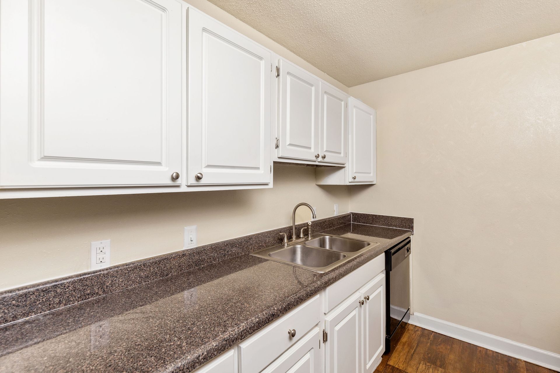 A kitchen with white cabinets, dark speckled countertops, a double basin sink, and a black dishwasher on wooden flooring.