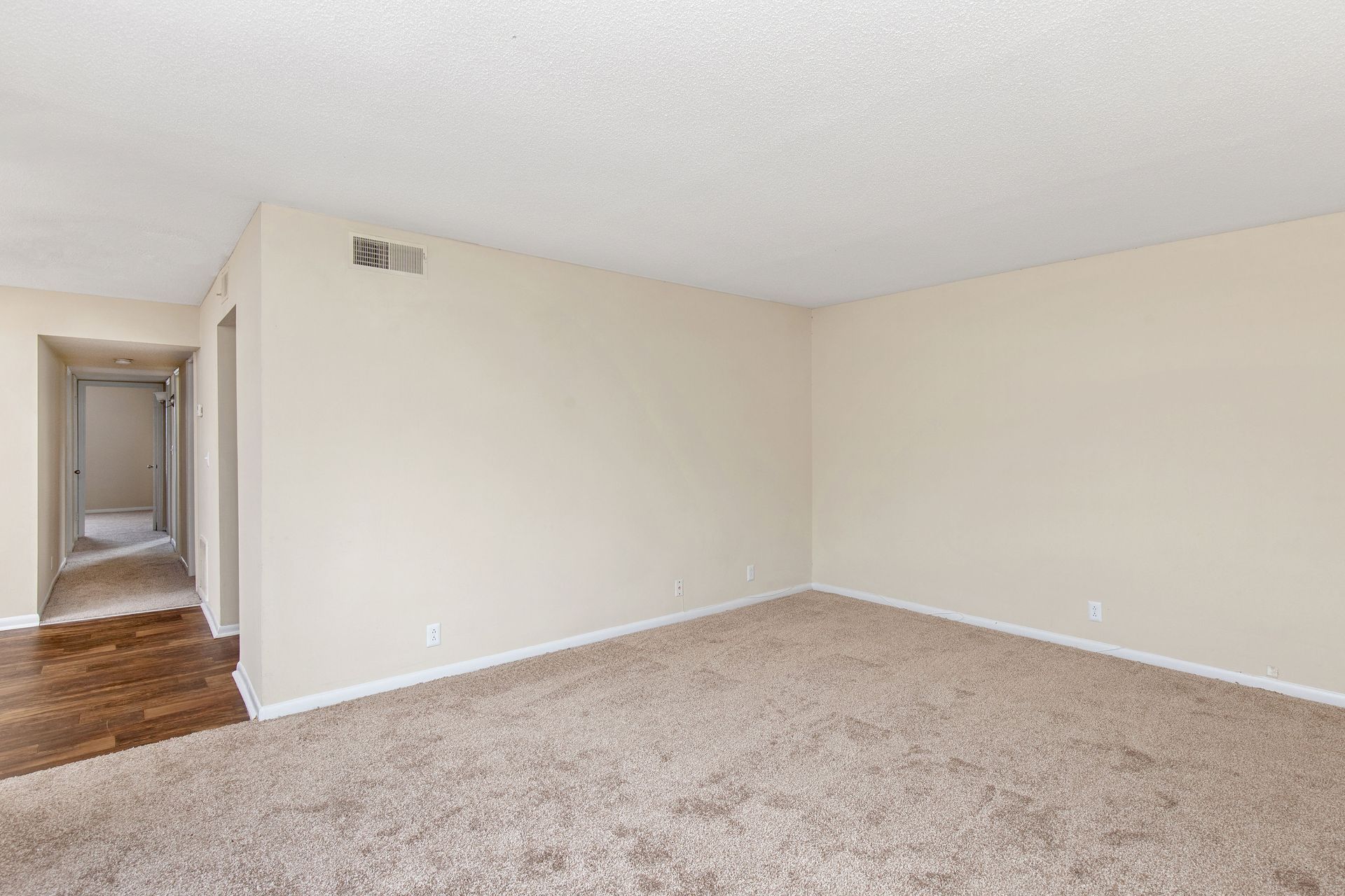An empty, beige-carpeted room with plain off-white walls, leading into a hallway with hardwood-style flooring.