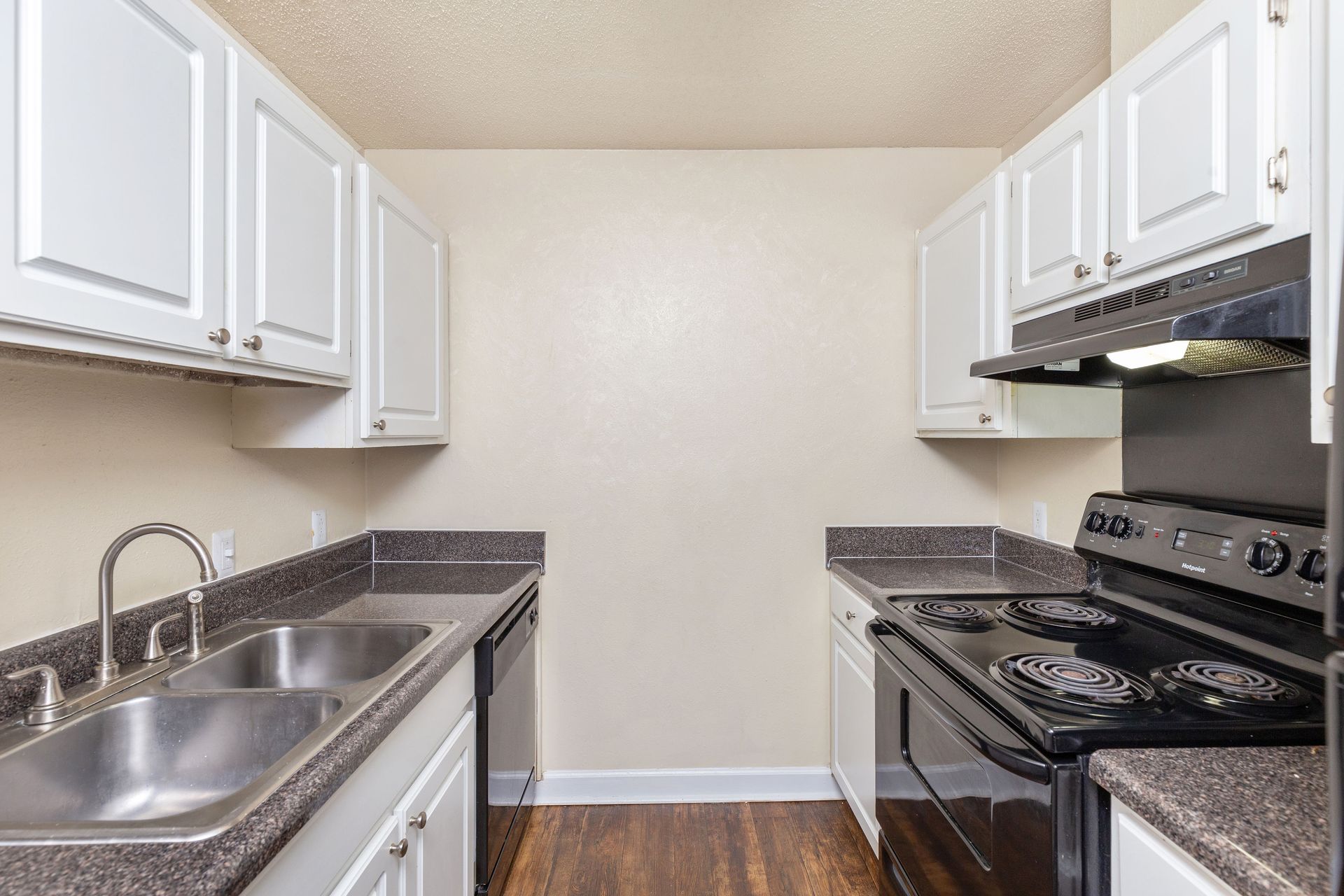 A galley-style kitchen with white cabinets, dark countertops, a double sink, a black stove, and brown wood-look flooring.