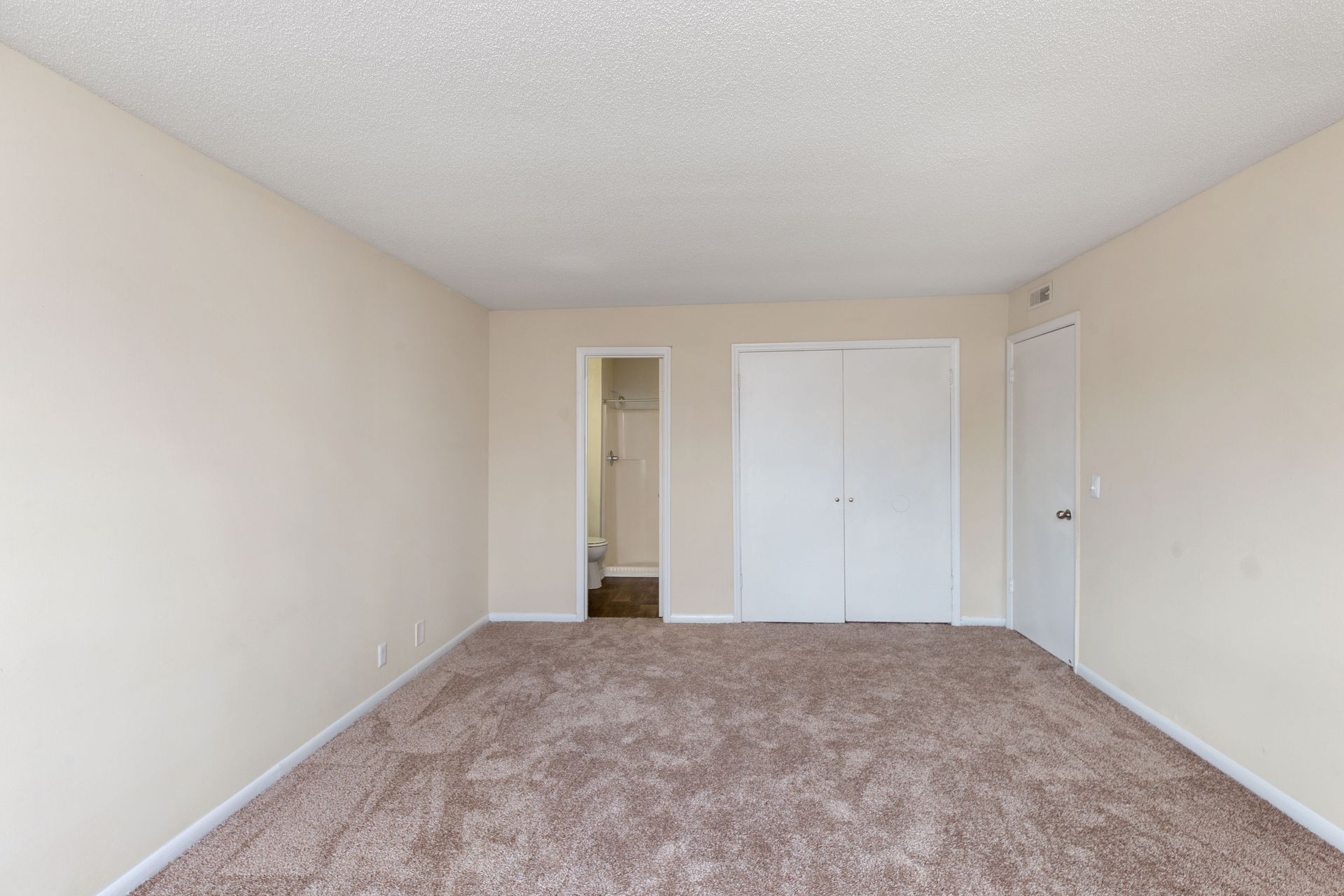 An unfurnished room with beige walls, brown carpet, a closet with white double doors, and a doorway leading to a bathroom.