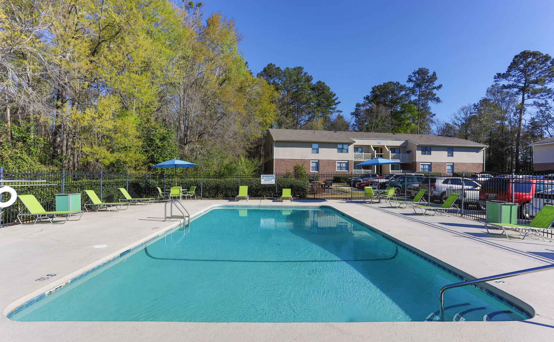 An outdoor rectangular swimming pool surrounded by a concrete deck with lounge chairs and apartment buildings behind.