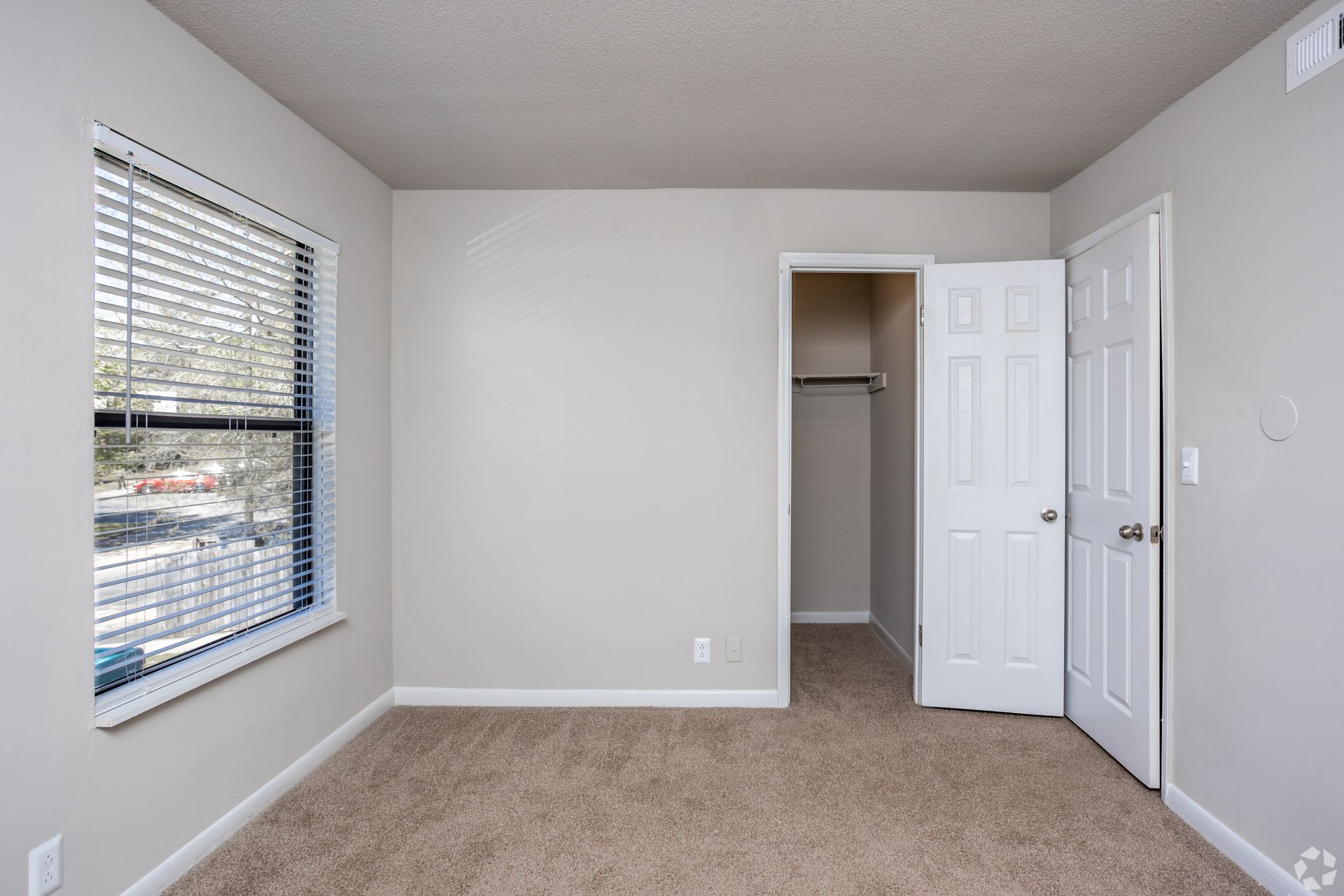 An empty bedroom with light gray walls, beige carpet, a window with blinds, and two white doors, one leading to a closet.