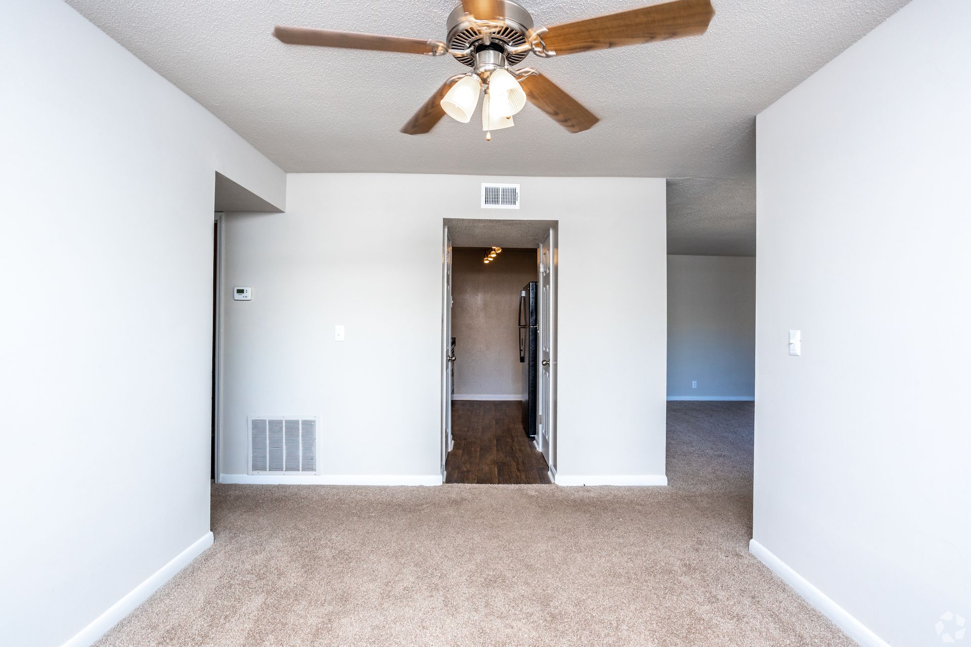 A carpeted room with beige walls, a ceiling fan, and a doorway leading to a kitchen with dark flooring.