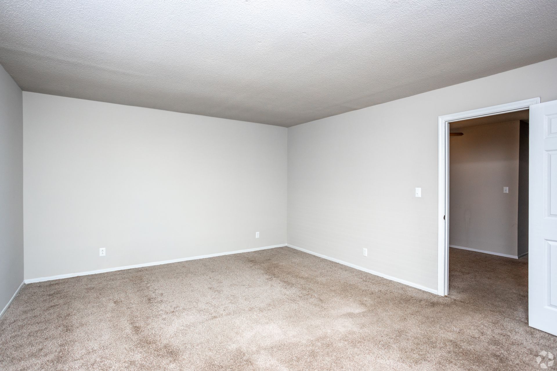 An empty room with light gray walls, textured white ceiling, and beige carpeting, featuring an open doorway on the right.