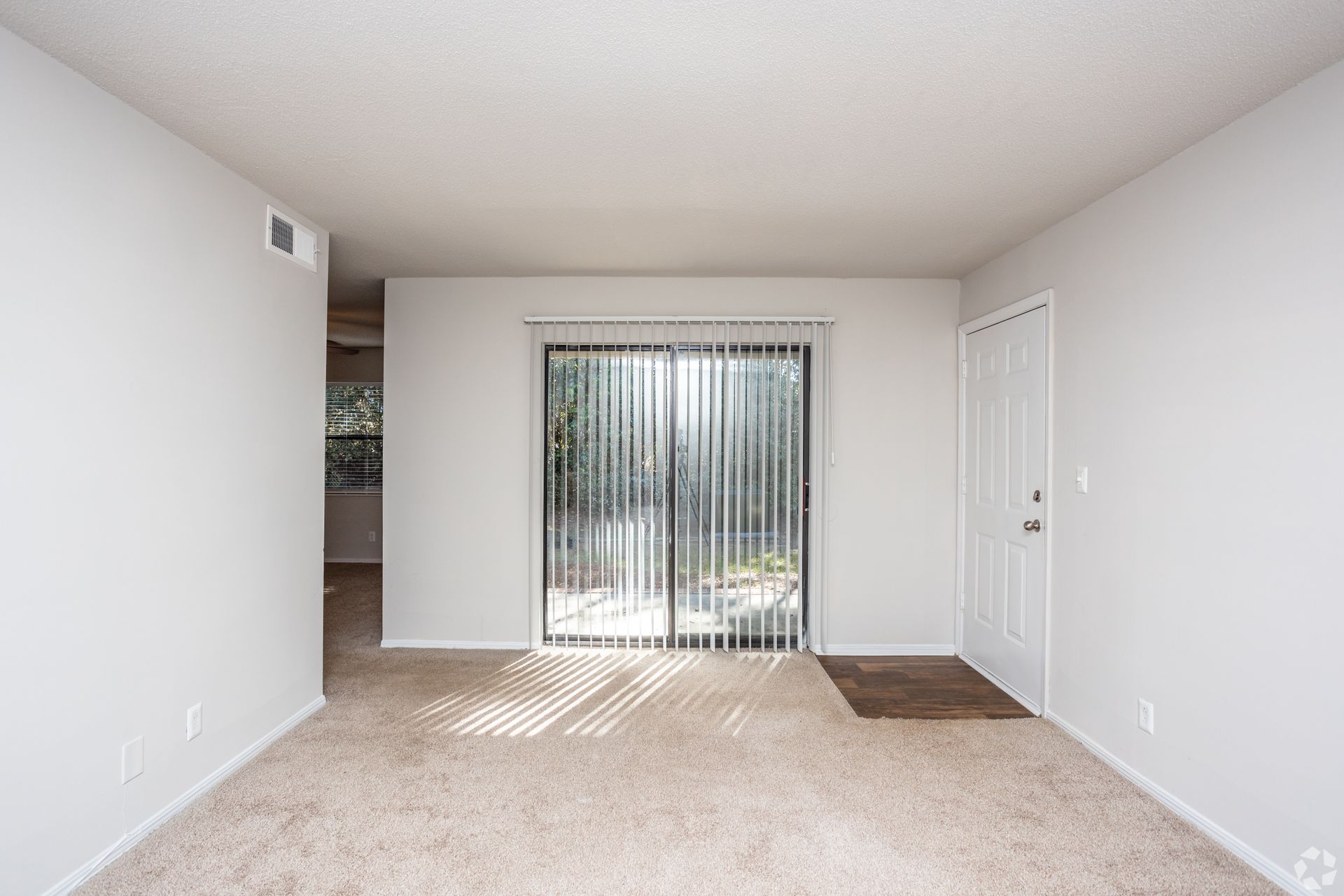An empty, neutrally colored living room with beige carpeting, a white door, and a sliding glass door with vertical blinds.