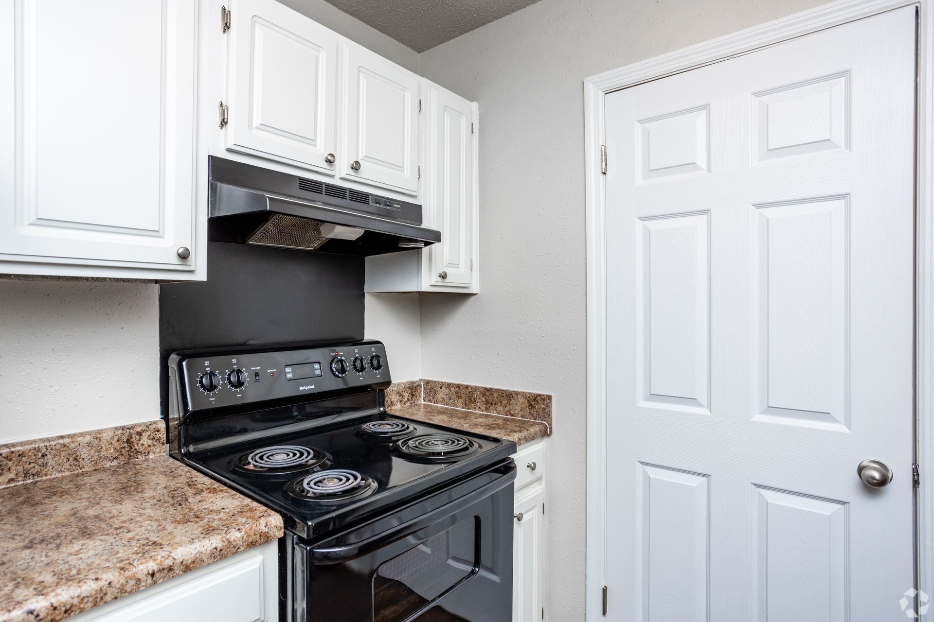 A kitchen with white cabinets, a black electric stove, a brown speckled countertop, and a white six-panel door.