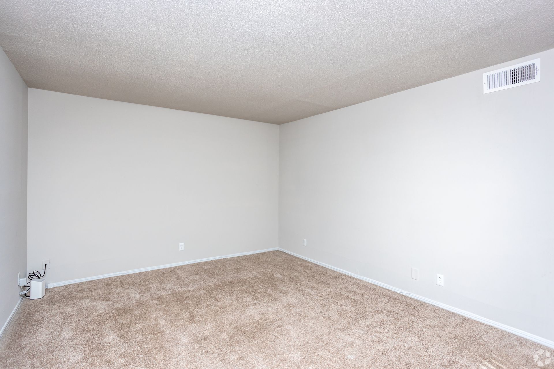 An empty room with light gray walls, beige carpeting, a popcorn ceiling, and a vent on the upper wall.