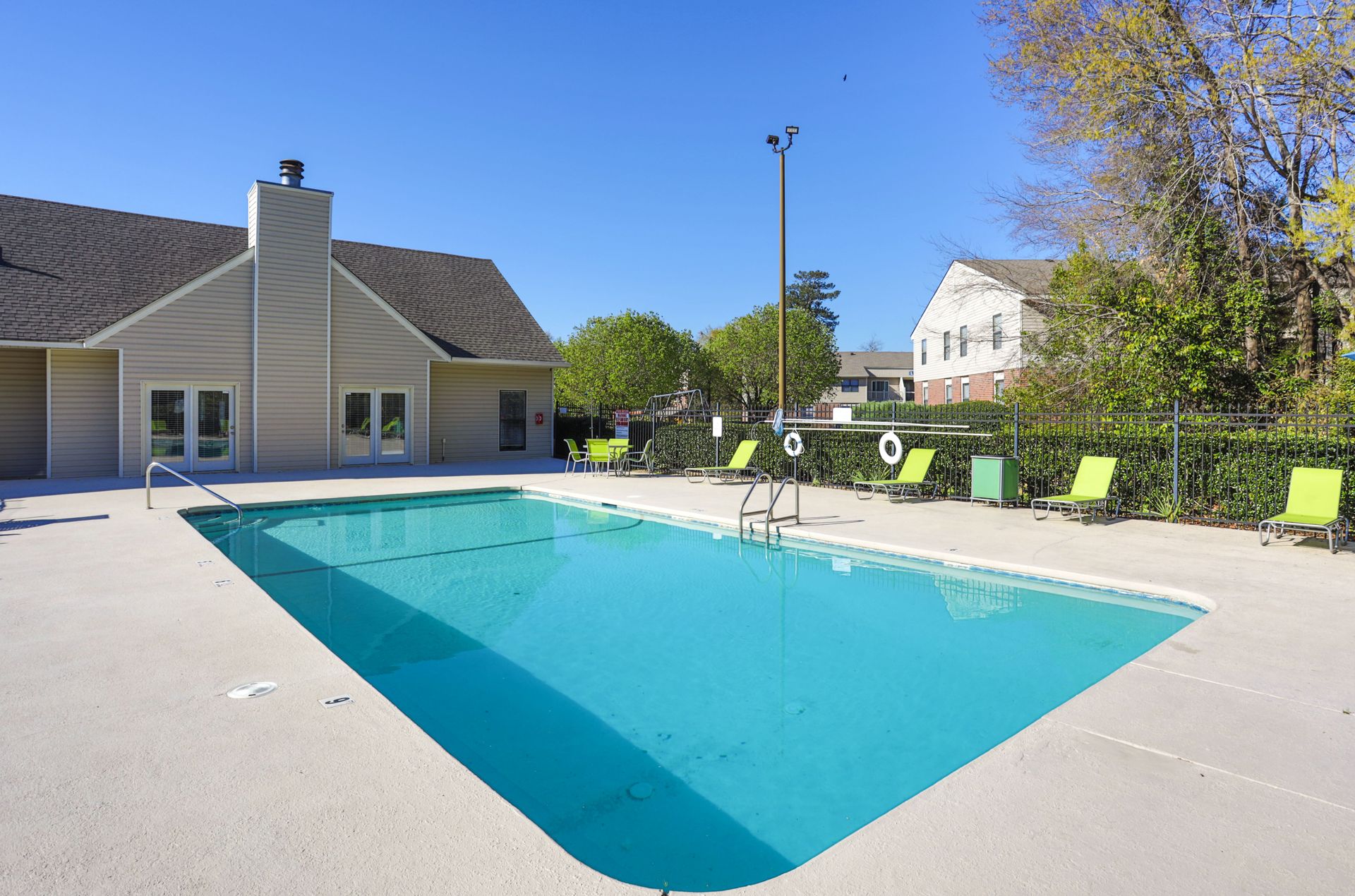A rectangular swimming pool with bright blue water sits in a concrete deck next to a beige building under a clear sky.