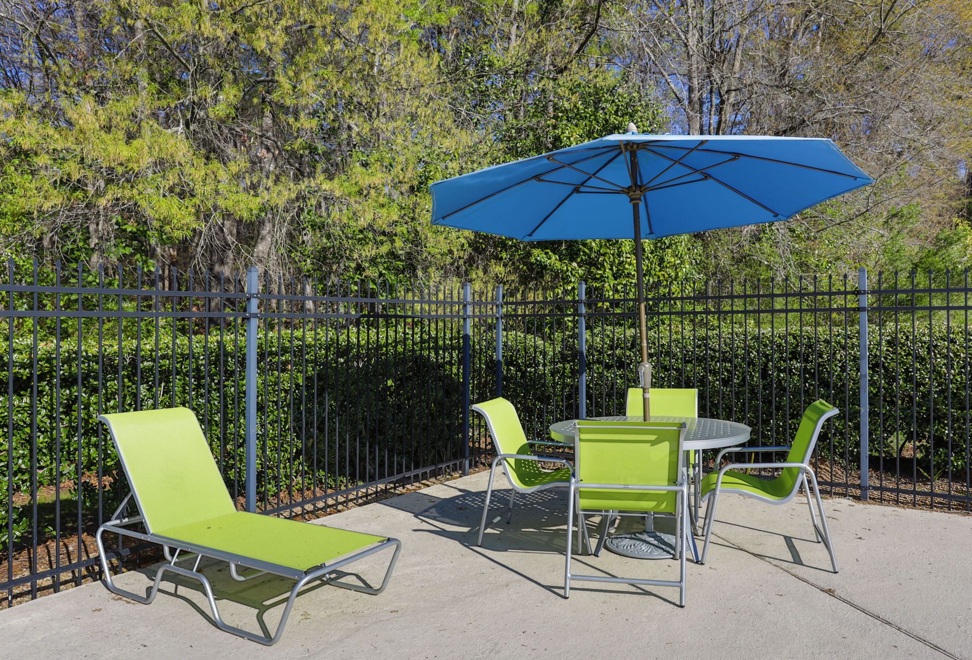 A bright blue patio umbrella over a dining set with four lime green chairs, beside a lime green lounge chair on a patio.