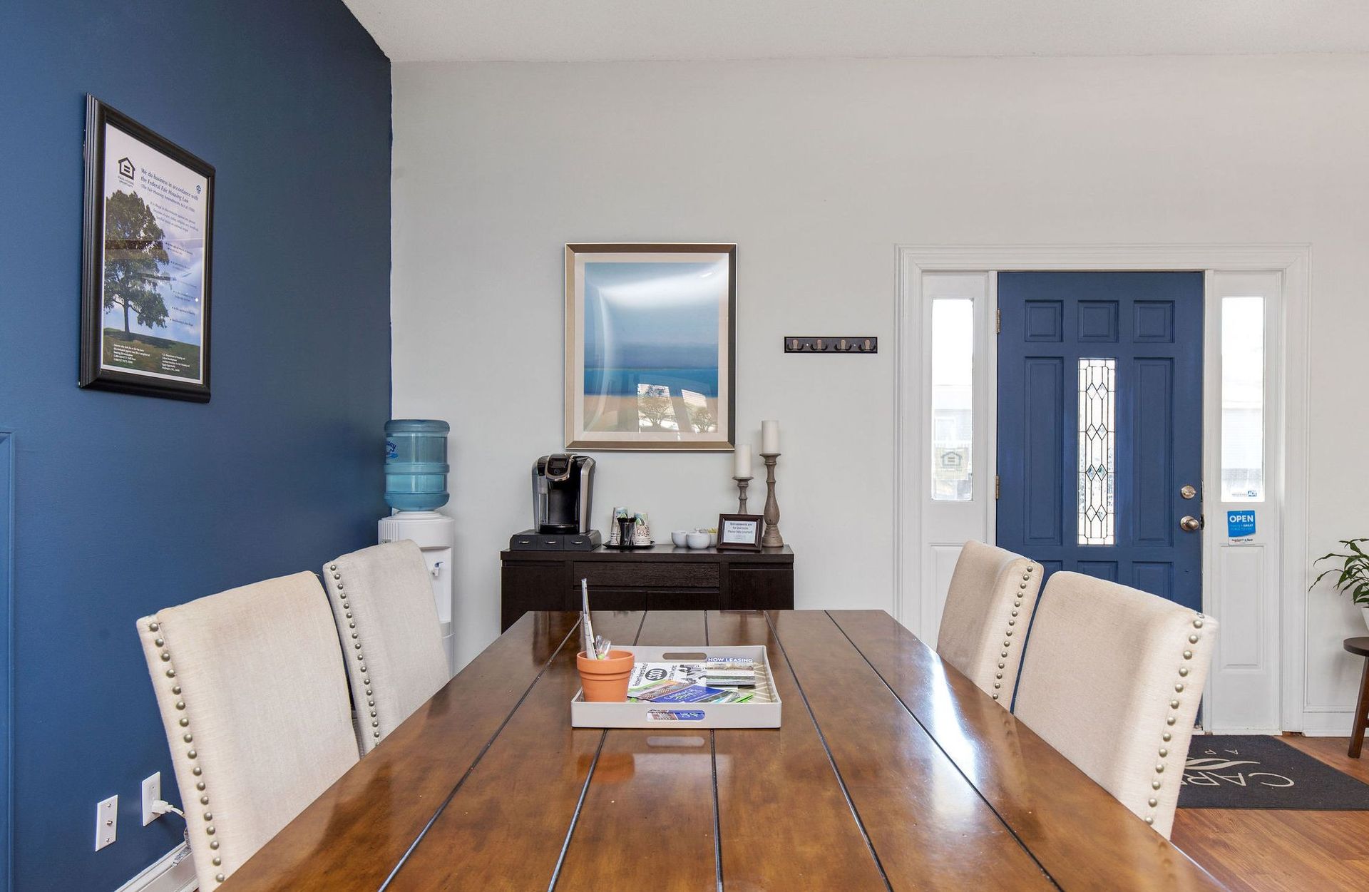 A wooden dining table with upholstered chairs sits in front of a blue accent wall and an entry door.