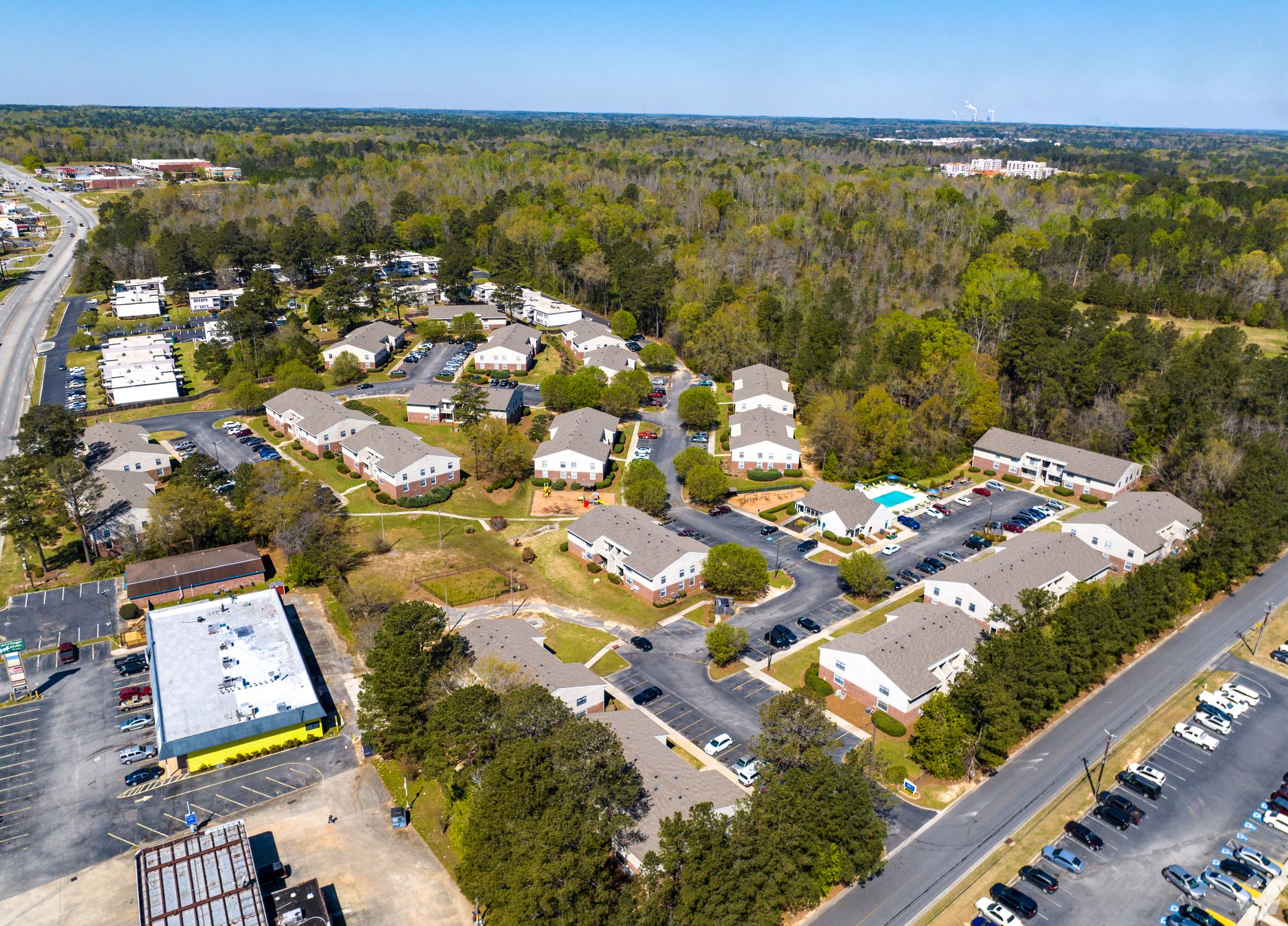 Aerial view of a residential apartment complex surrounded by trees, featuring multiple buildings, parking, and a pool.