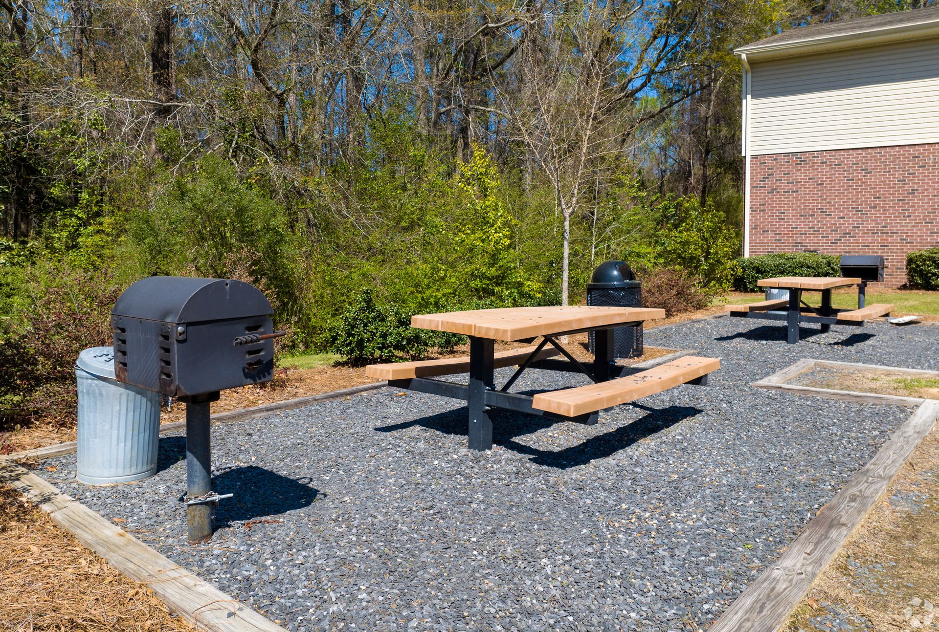 Two picnic tables and two metal grills on a gravel pad next to a brick building and a wooded area.