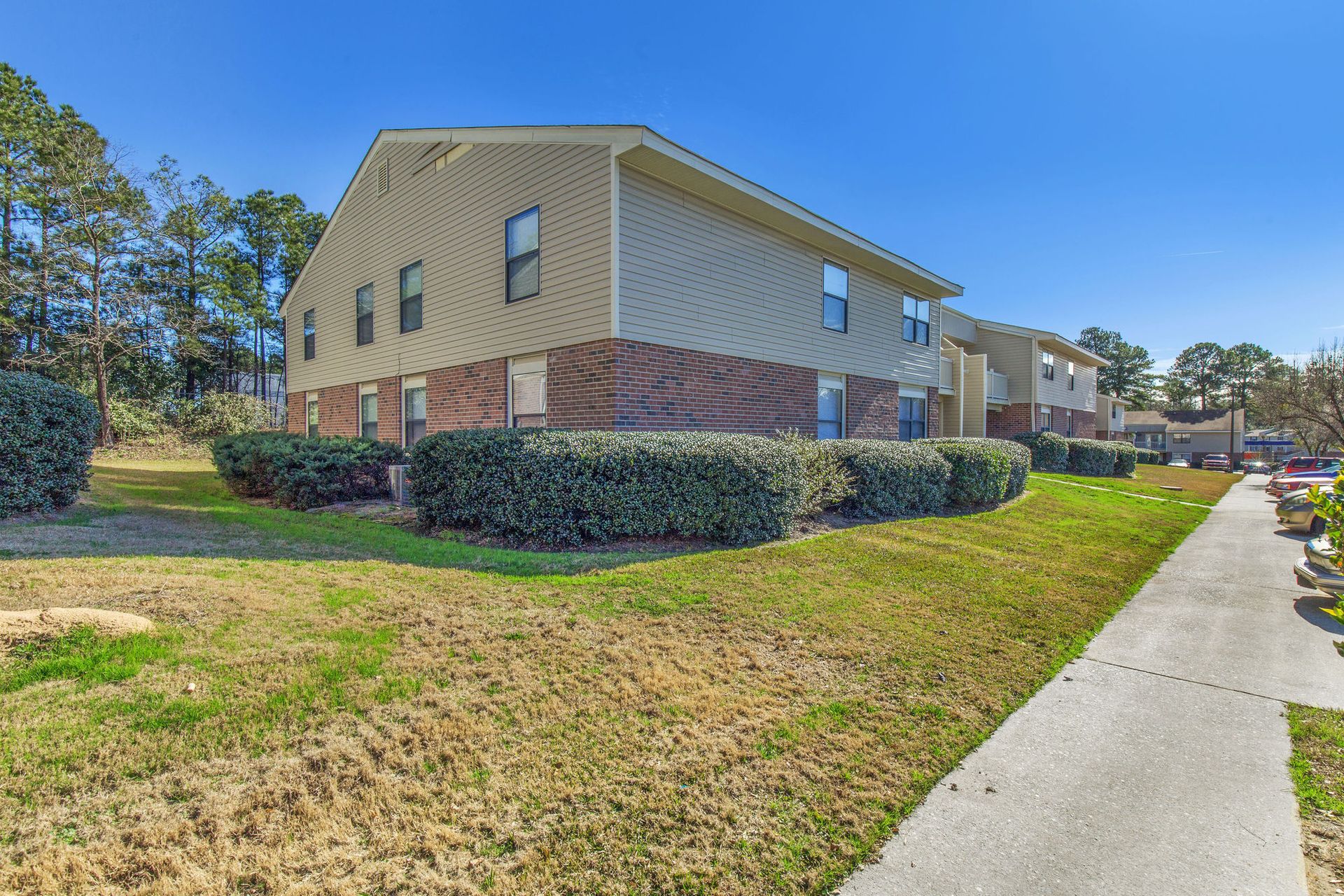 A two-story apartment building with tan siding and brick accents, surrounded by landscaping under a clear blue sky.