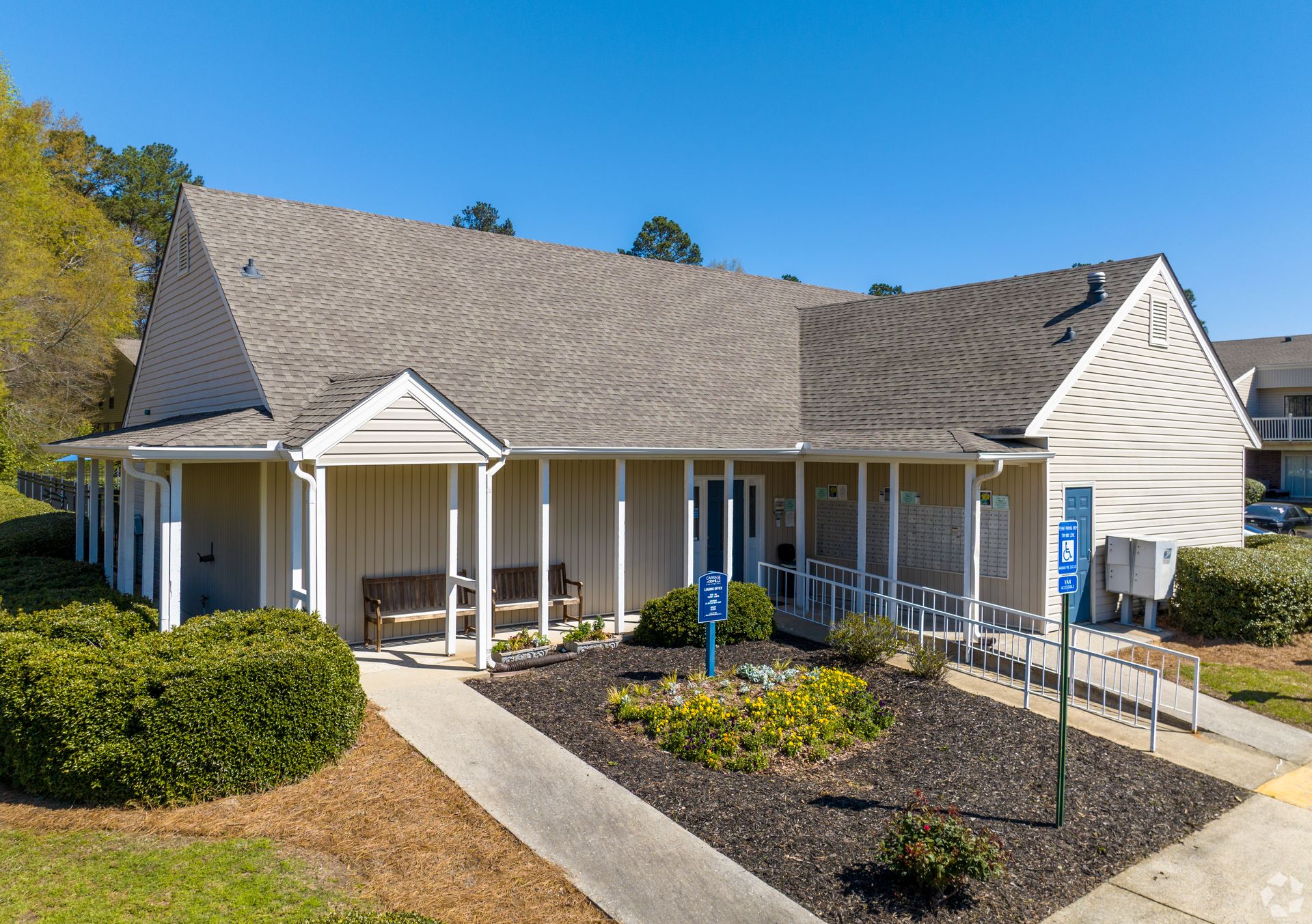 A tan, single-story community center building with a gray shingled roof, a front porch, and handicap parking signs.