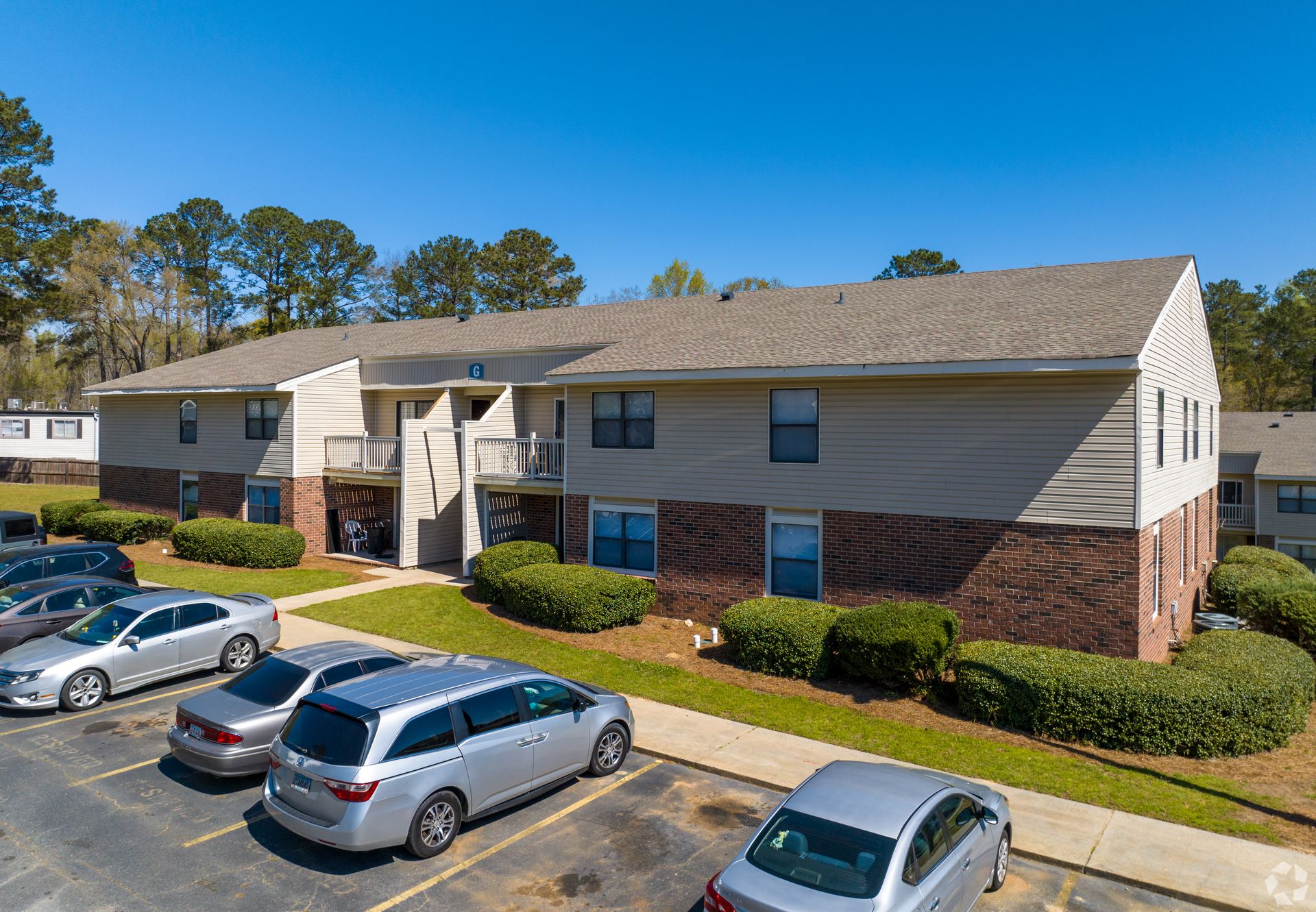 An exterior view of a two-story apartment building with tan siding, brick accents, and cars parked in front.