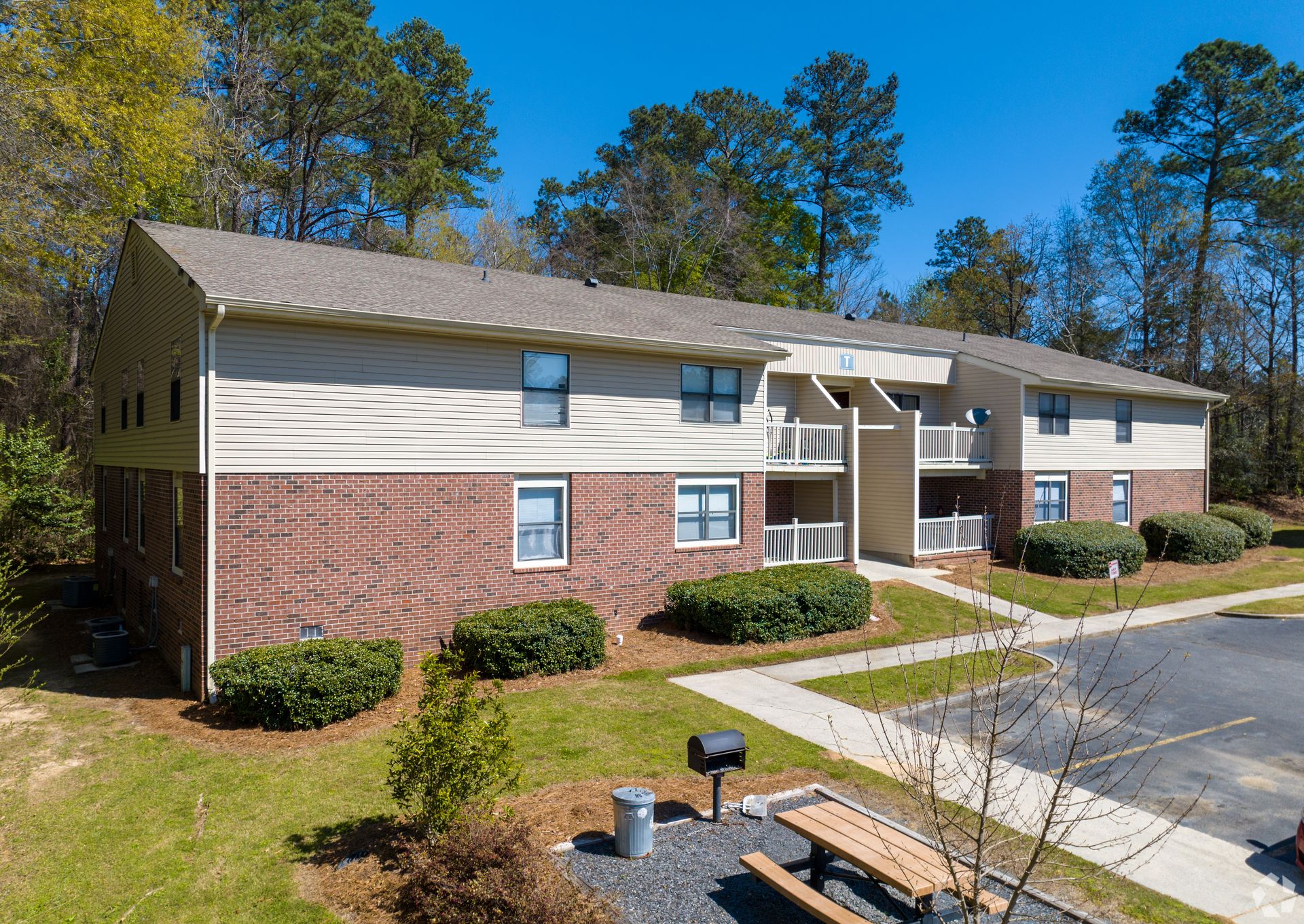 A two-story apartment building with beige siding and brick ground level, featuring a sidewalk and picnic area in front.