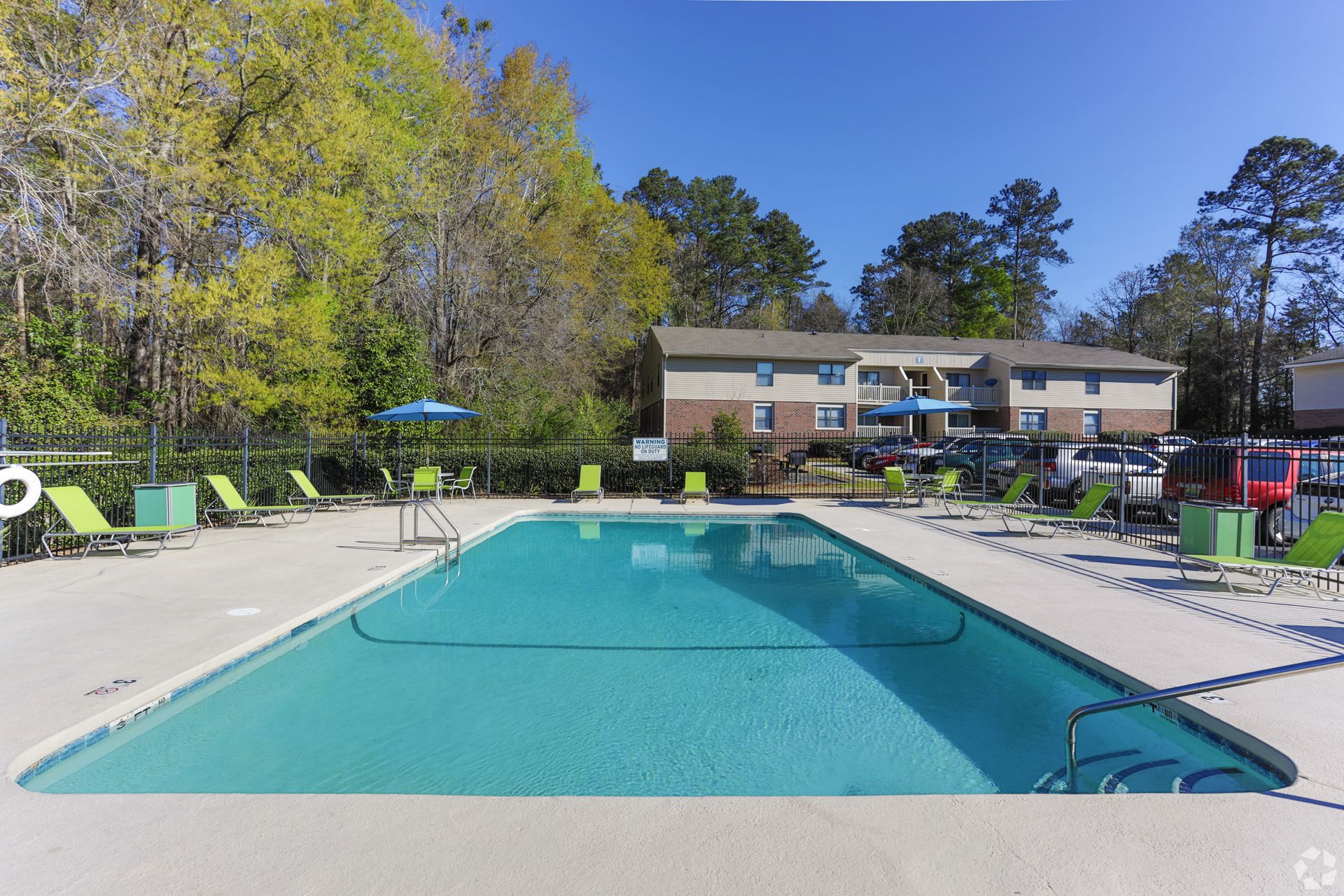 An outdoor swimming pool surrounded by a concrete deck with lounge chairs, trees, and an apartment building in the back.