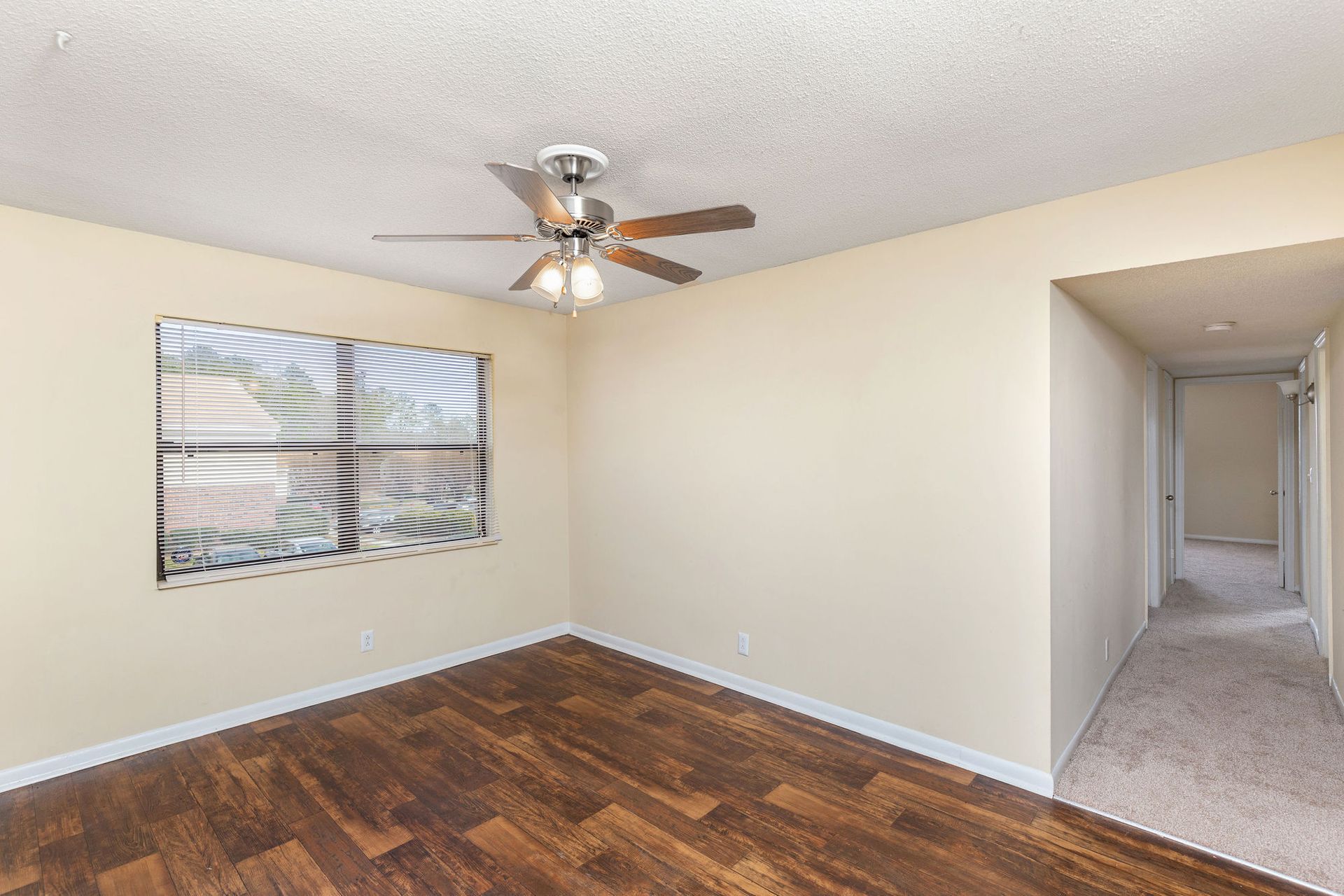 An empty room with beige walls, wood floors, a ceiling fan, a window with blinds, and an open doorway to a hallway.