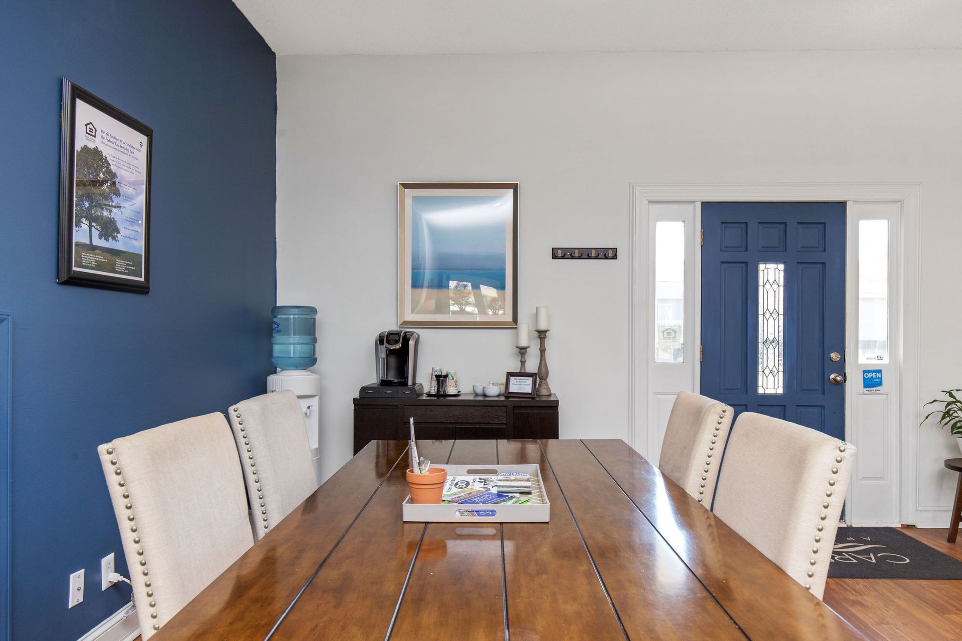 Dining table and chairs in a lobby with a blue accent wall, framed art, coffee station, and blue front door.