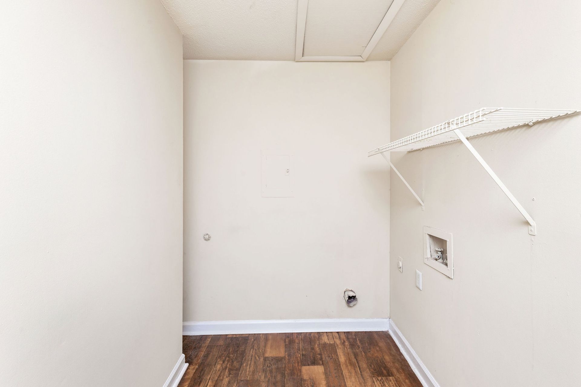 An empty laundry room with light-colored walls, wood flooring, a wire shelf, and utility hookups for a washer and dryer.