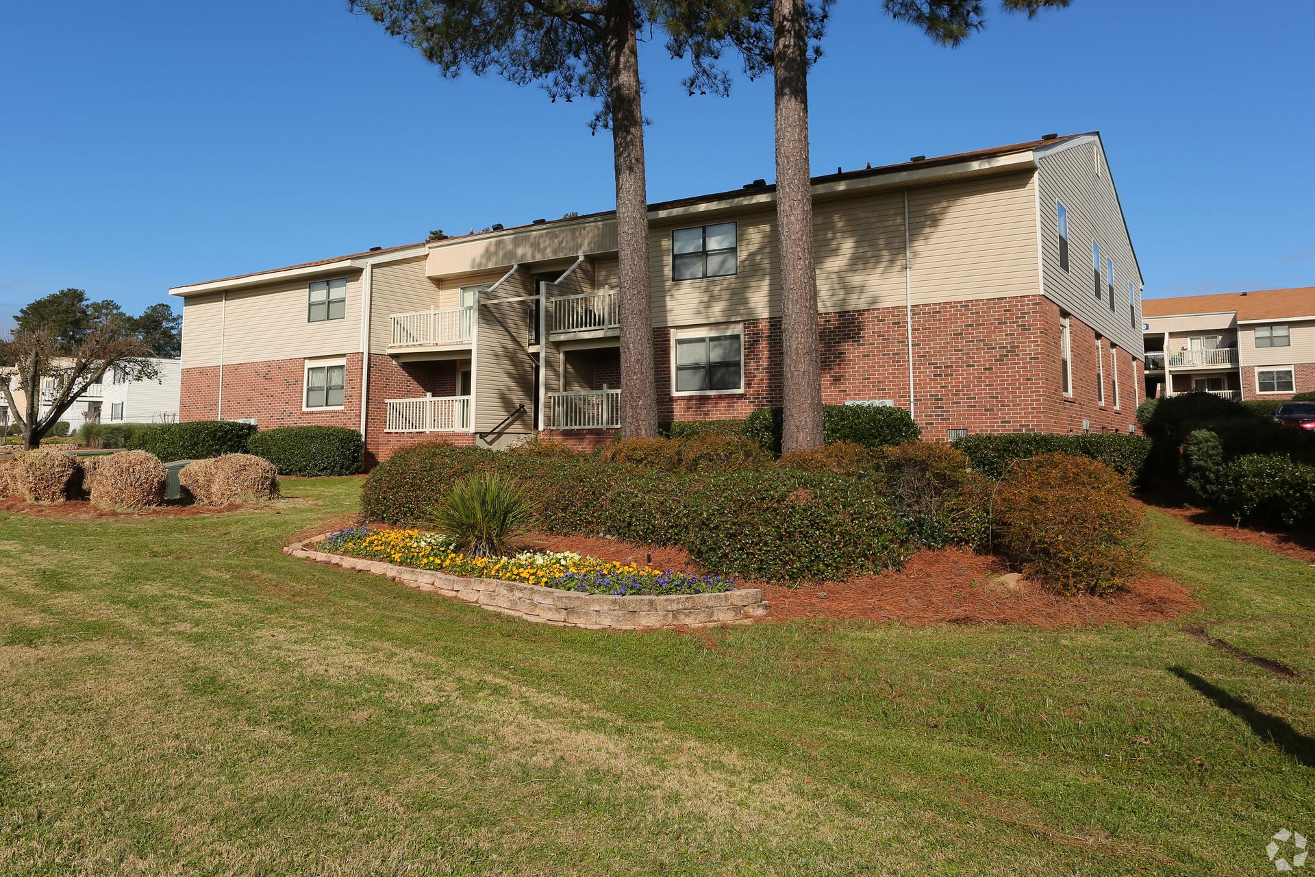 Two-story apartment building with brick and beige siding, surrounded by a lawn and manicured shrubbery under a blue sky.