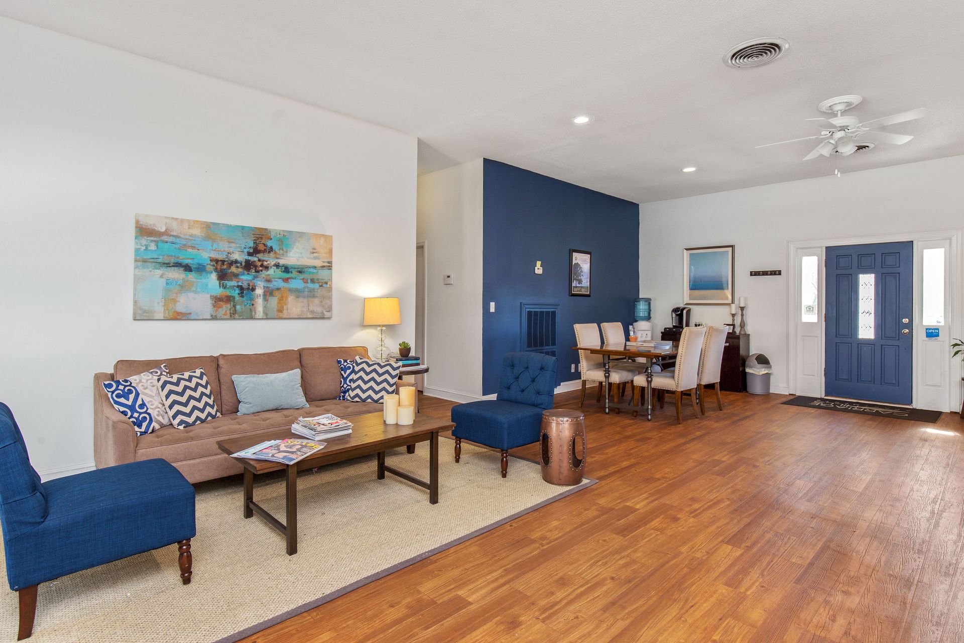 A living room with a beige sofa, blue accent chairs, a wooden coffee table, and a dining area against a navy feature wall.
