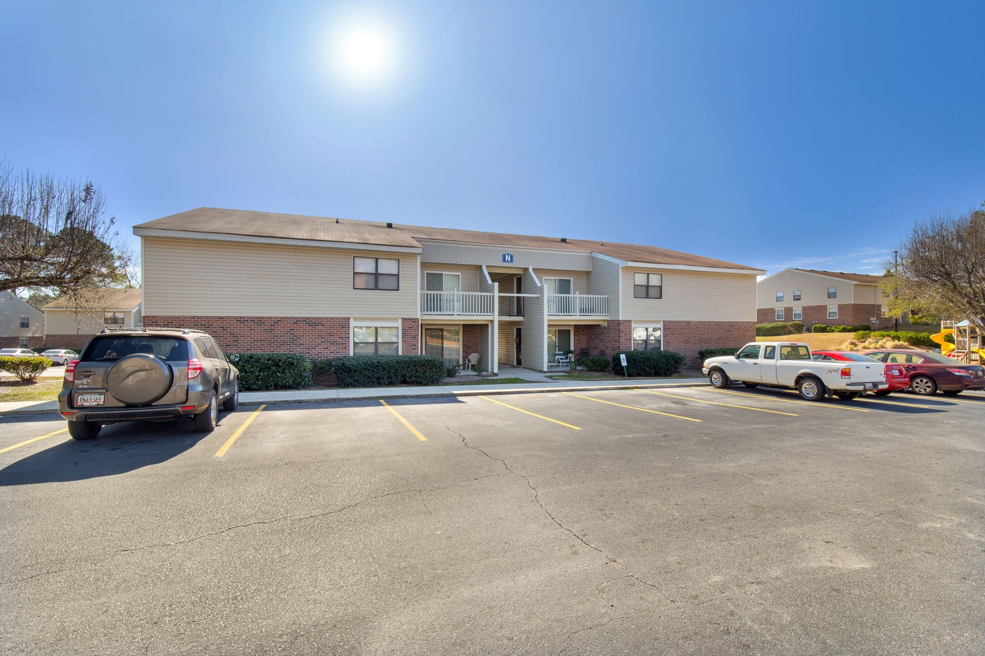 A two-story apartment building with tan siding and brick lower levels, viewed from a parking lot on a sunny day.