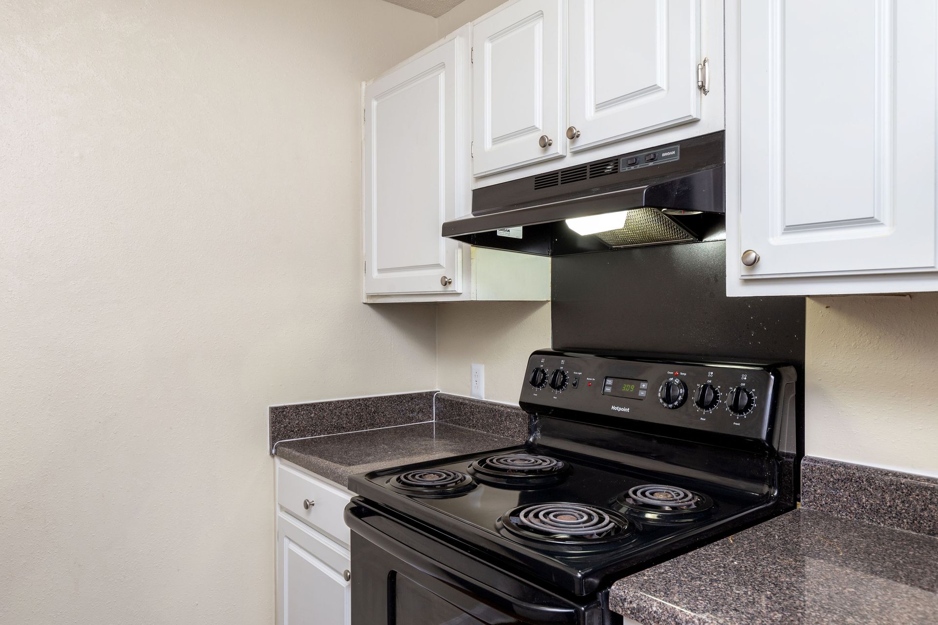 Kitchen view featuring a black stove, range hood, and speckled brown countertops with white cabinets above.