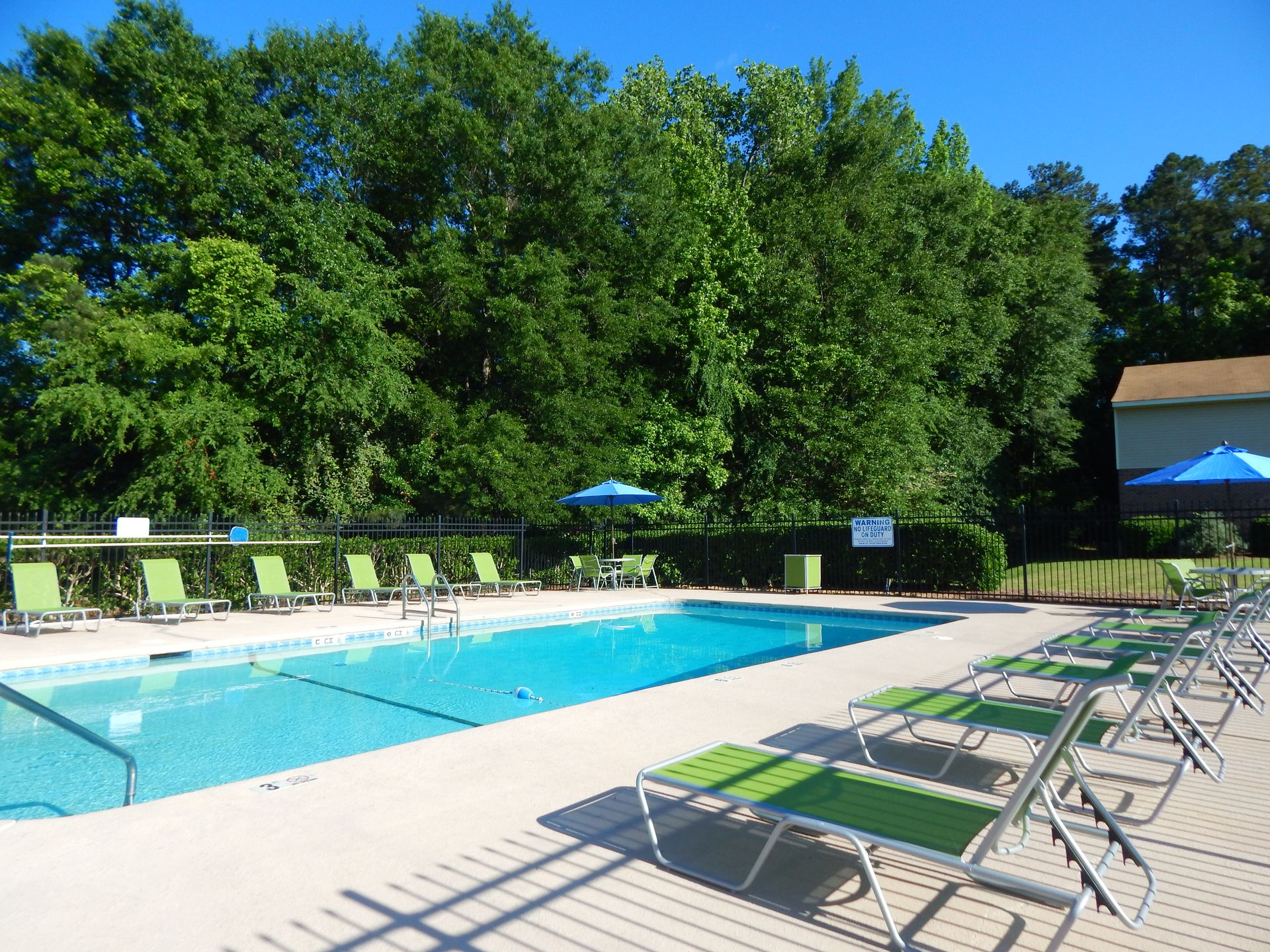 Outdoor swimming pool surrounded by green lounge chairs and blue umbrellas, set against a backdrop of dense trees.