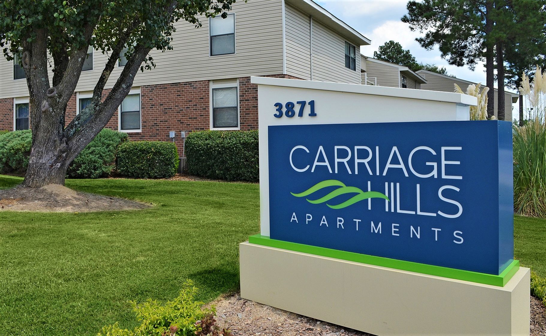 A blue sign for Carriage Hills Apartments numbered 3871, located in front of a multi-story residential building and tree.