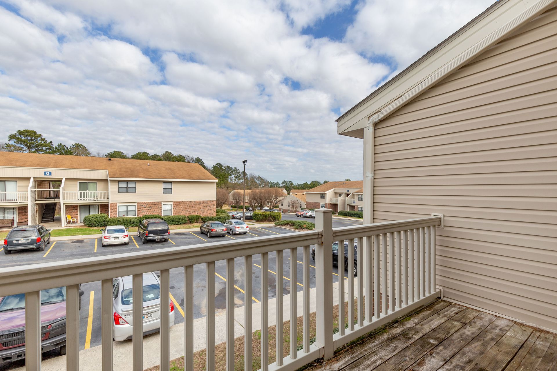 A view from a wooden balcony overlooking a parking lot with several parked cars and apartment buildings under a cloudy sky.