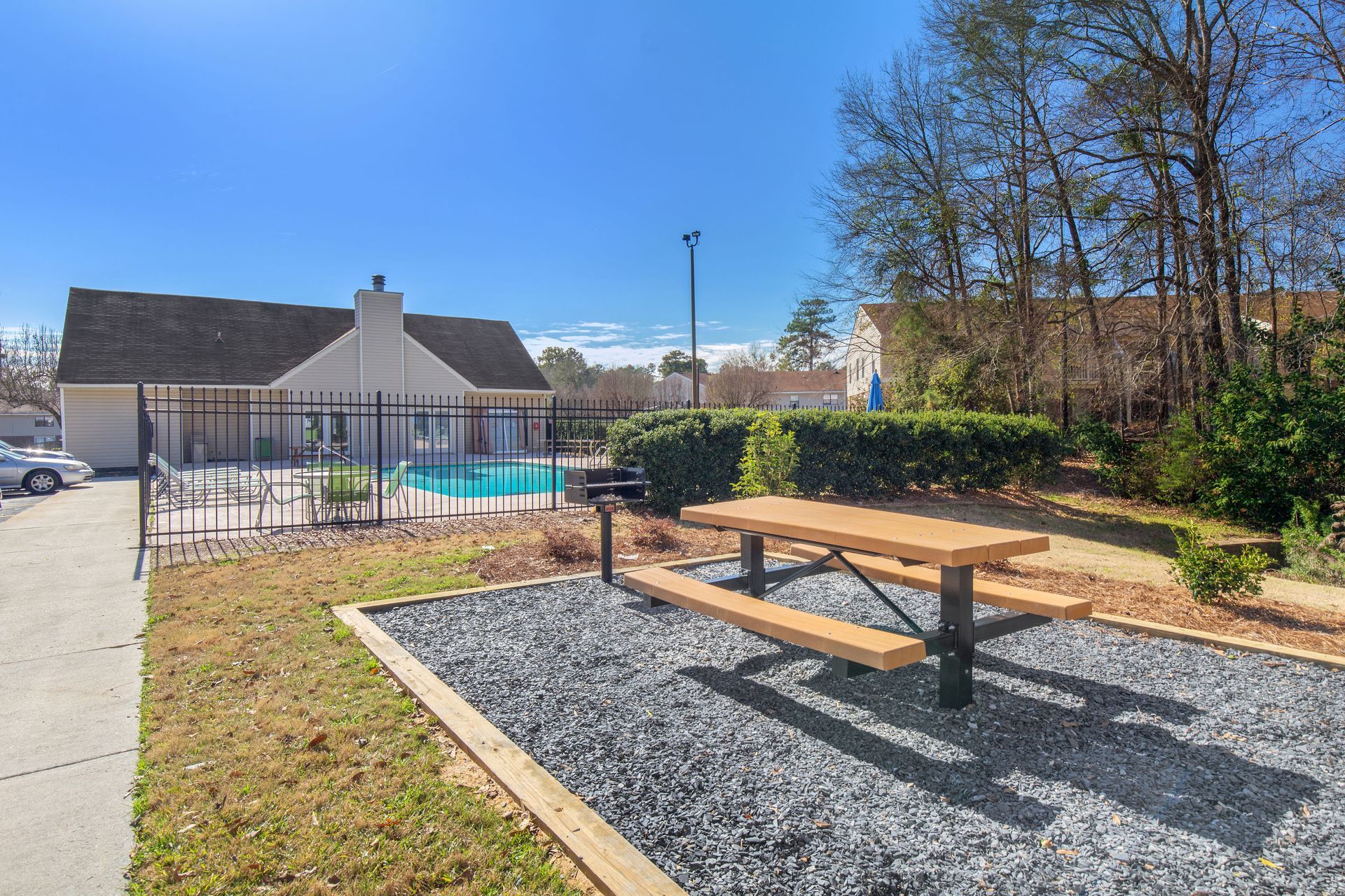 A picnic table on a gravel pad with a grill next to a fenced-in swimming pool area near trees under a clear blue sky.