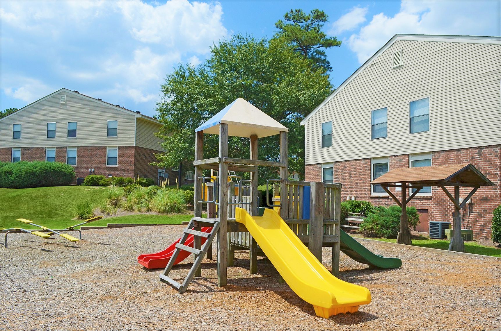 A playground with a multi-level climbing structure, yellow and red slides, and a nearby wooden shelter between buildings.