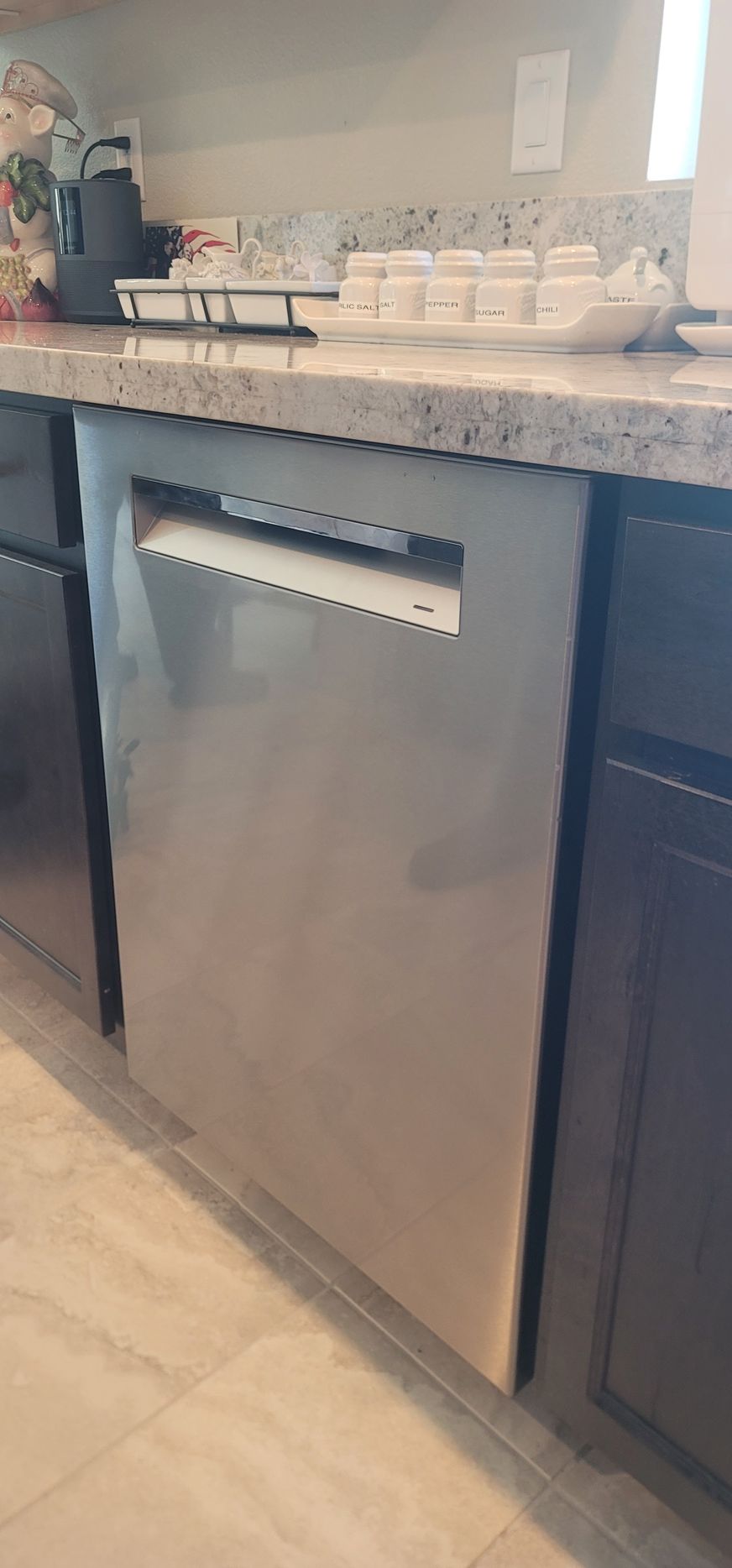 Stainless steel dishwasher beneath a granite kitchen counter with dark cabinets and tile flooring