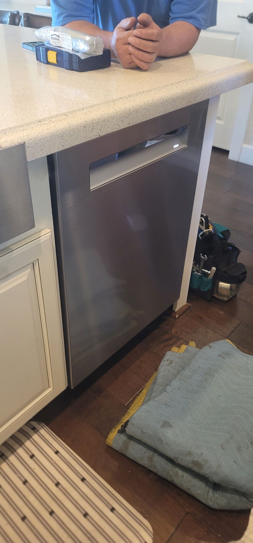 Person leaning on kitchen counter above a built-in dishwasher with cleaning items on the floor