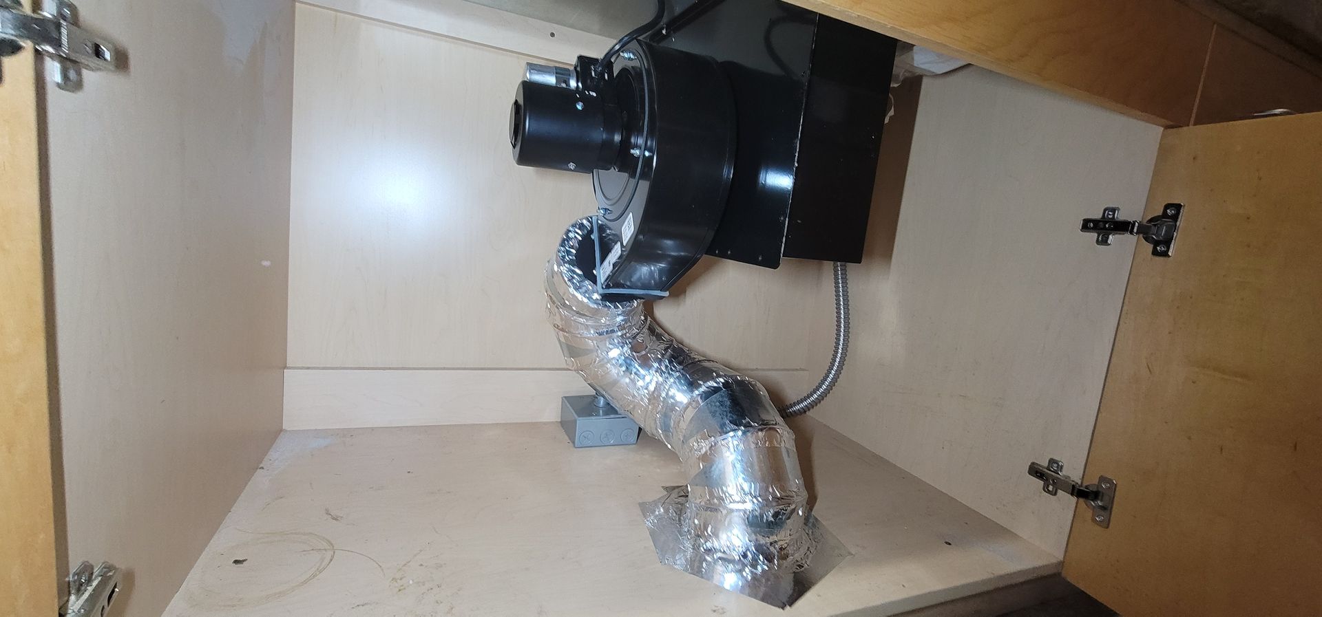 Under-sink cabinet with a black vent fan and shiny silver ducting installed inside.