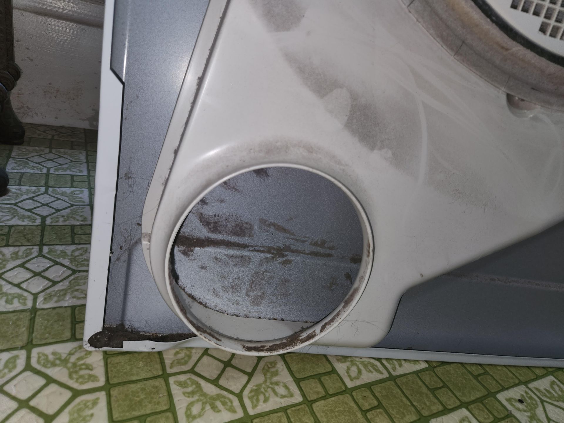 Close-up of a white appliance with a round vent and scuffed lower panel on tiled floor