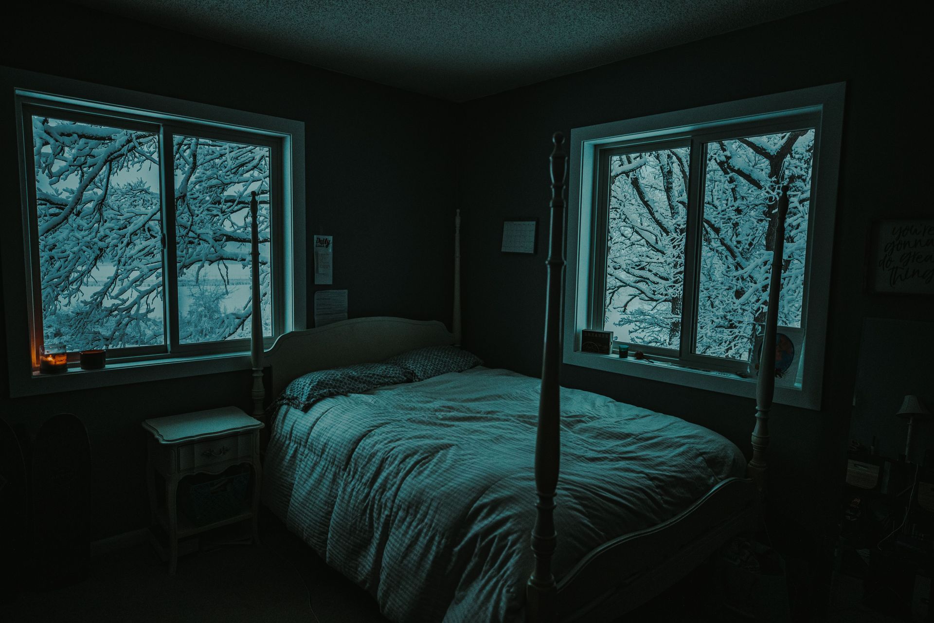 Dark bedroom with a bed between two snow-covered windows and a teal glow.