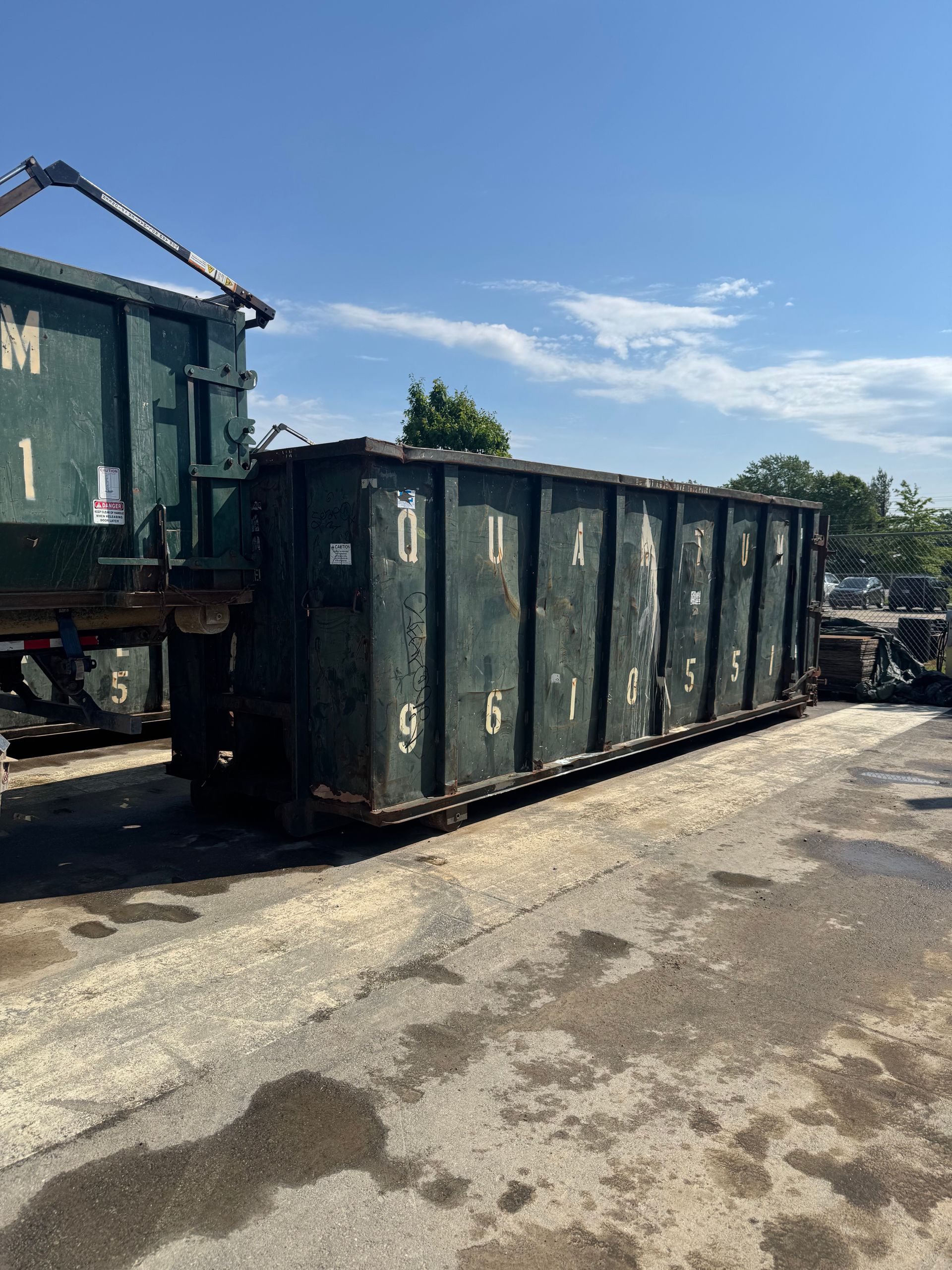 Row of dark green industrial dumpsters on a sunny paved lot beside a truck.