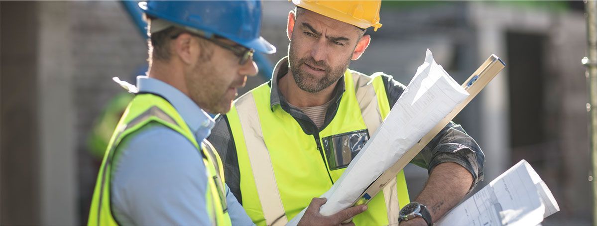 Two construction workers in hard hats and safety vests reviewing blueprints outdoors
