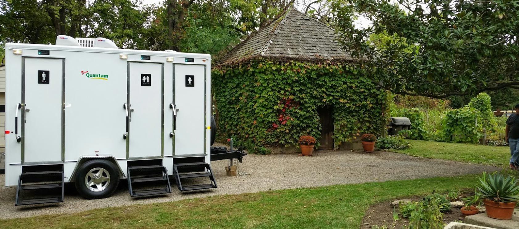 White portable restroom trailer on gravel driveway beside a hedge and thatched cottage in a garden