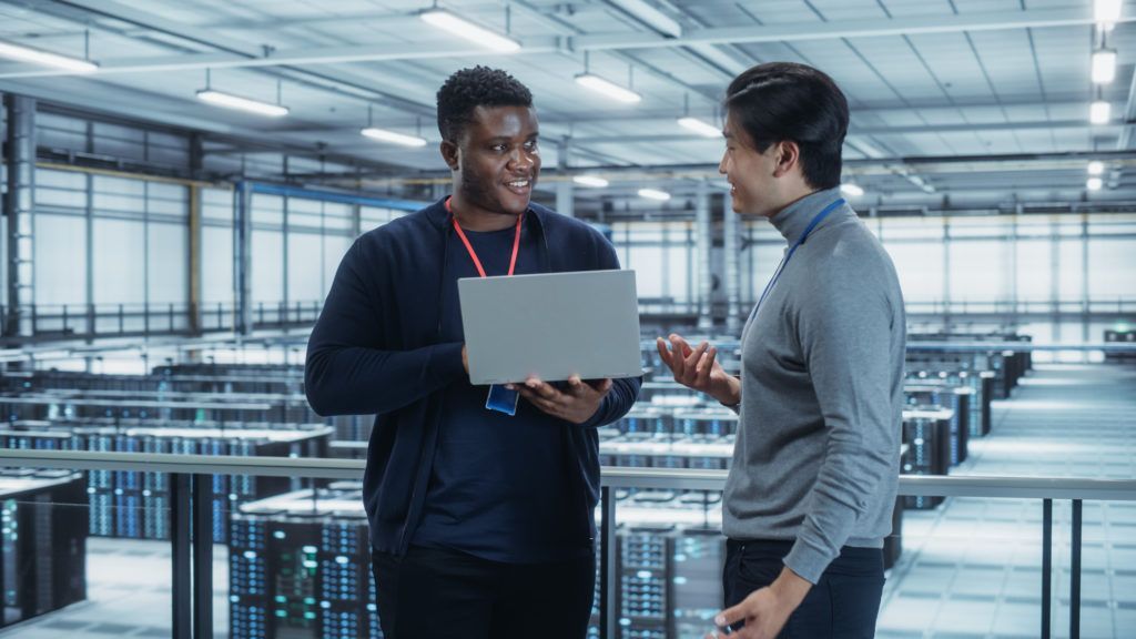 Duas pessoas em uma sala de servidores, uma delas segurando um laptop. Sorrindo e conversando, cercadas por racks de computadores.